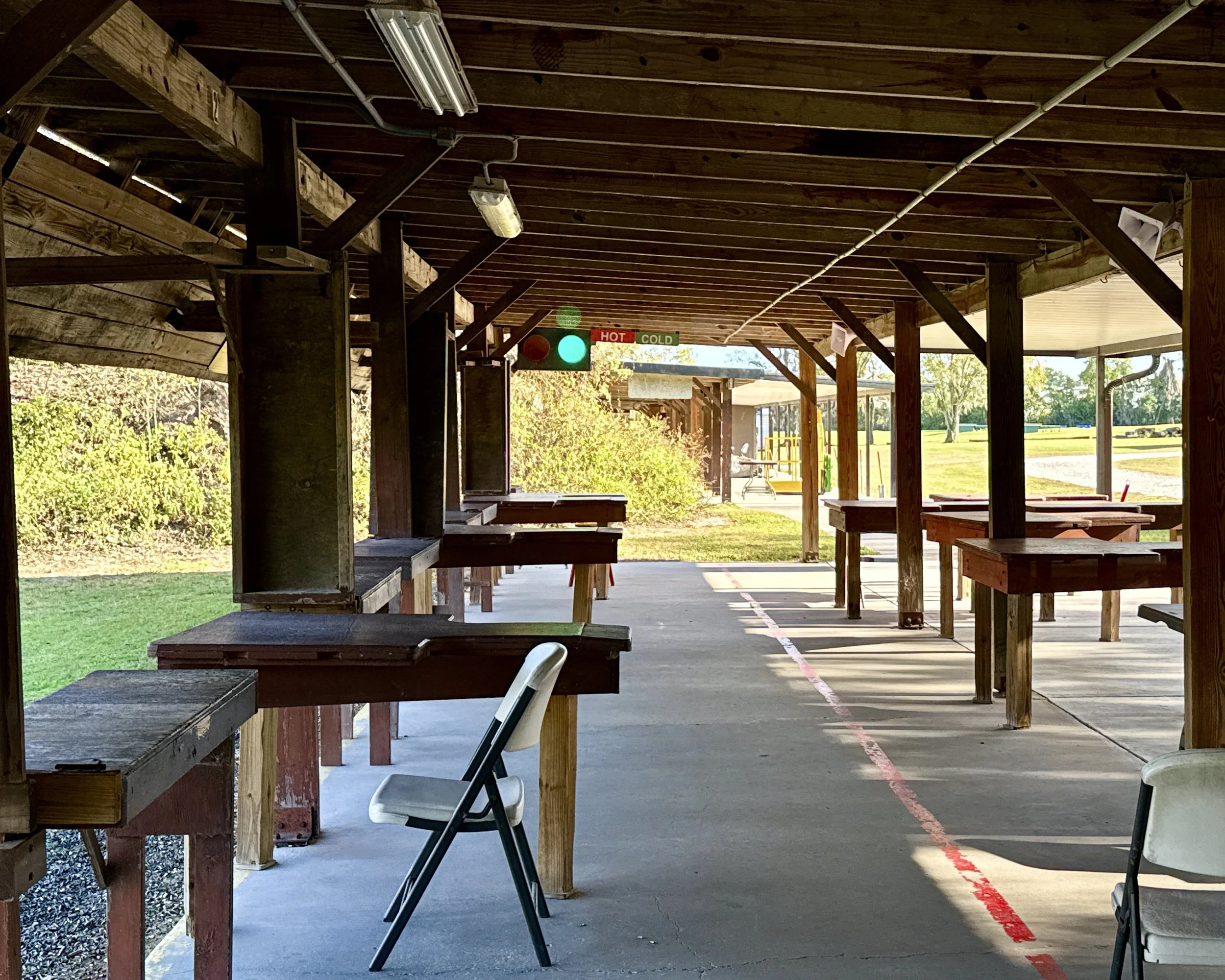 Empty outdoor shooting range with wooden booths, target stands, and a scoreboard with 'HOT' and 'COLD' signs. Sunlight filters through the open sides, and trees are visible in the background.
