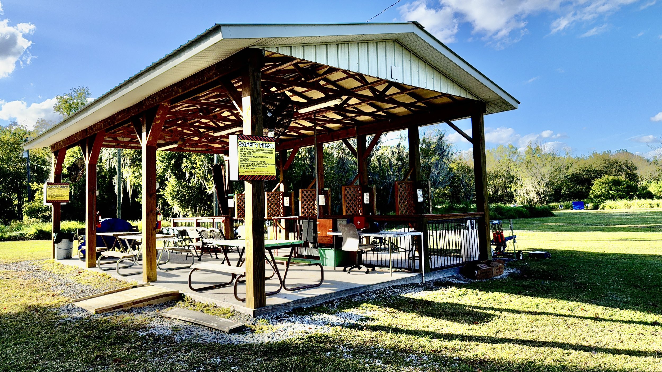 An outdoor covered 5-stand shooting structure with tables, chairs, and safety signs, surrounded by green grass and trees on a sunny day.