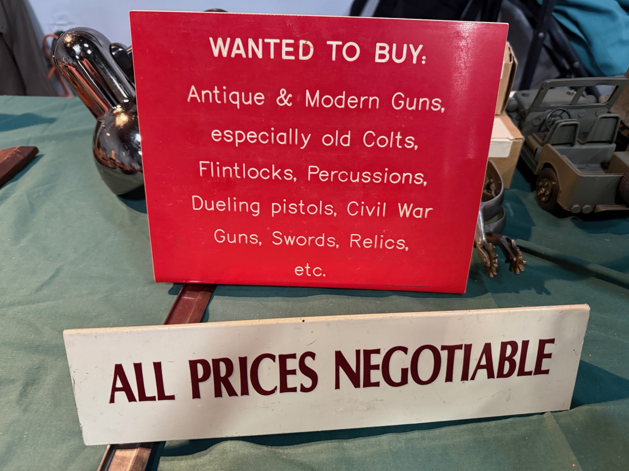 Red sign with white text reading "WANTED TO BUY: Antique & Modern Guns, especially old Colts, Flintlocks, Percussions, Dueling pistols, Civil War Guns, Swords, Relics, etc." next to a silver, reflective object on a table along with other vintage toy vehicles.