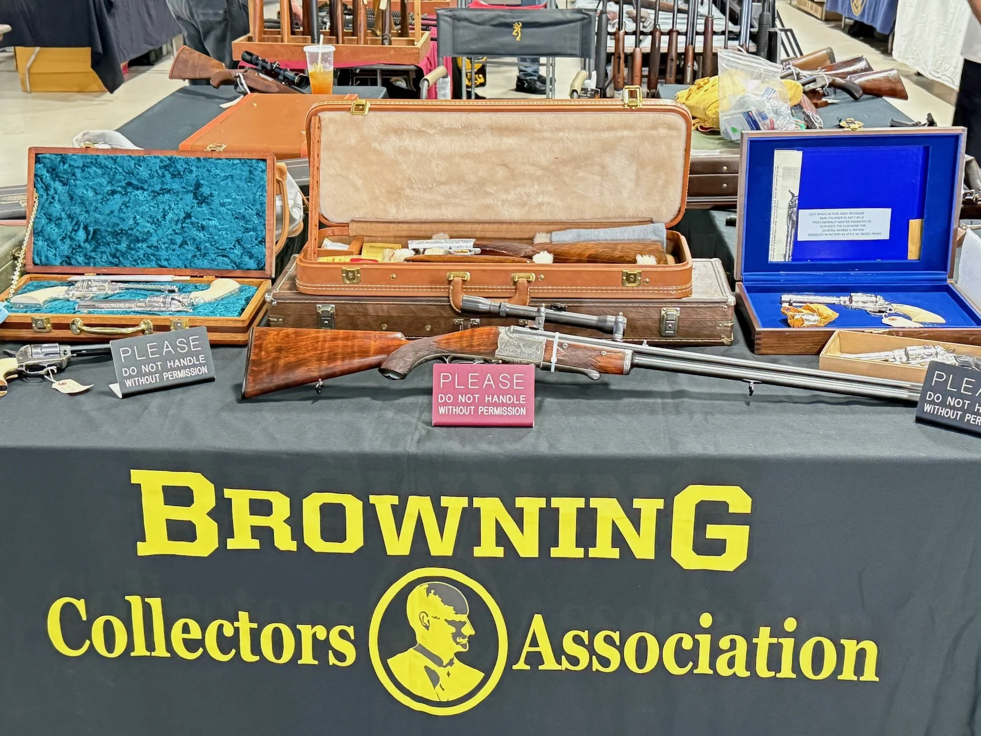 Table displaying collection of vintage firearms including rifles and guns in cases. Signage requests not to handle without permission. Cover of tablecloth reads "Browning Collectors Association" with an image of a man in profile.