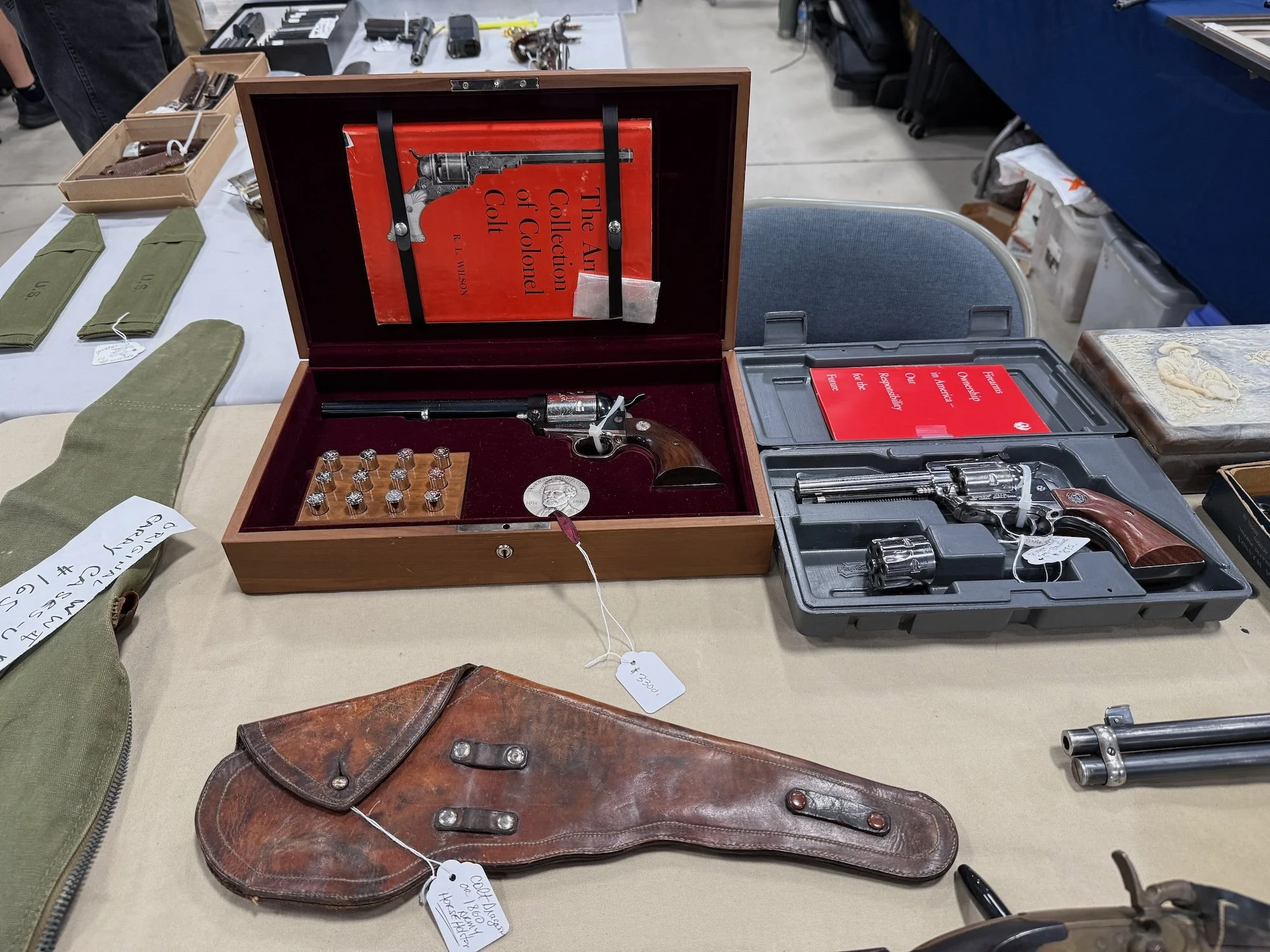 Display of vintage firearms including a revolver in a wooden case, another revolver in a plastic case, and a leather gun holster on a table at a gun show.