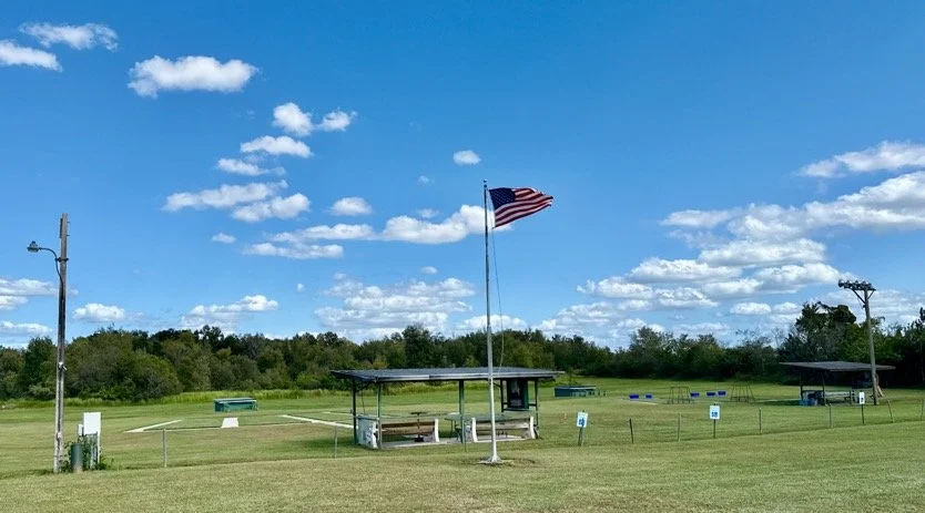 Trap field with a shelter, flagpole with American flag, and blue sky with scattered clouds.