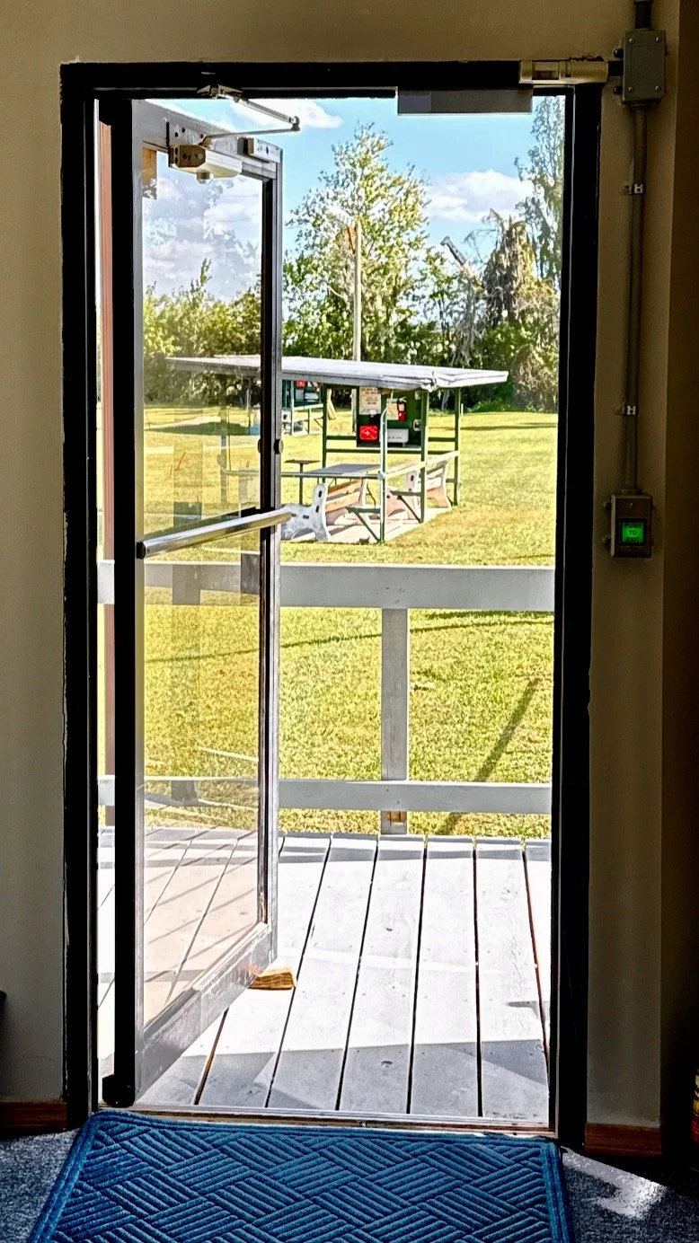 Open door leading to a grassy outdoor area with trap fields and a covered structure, bright blue sky with clouds.