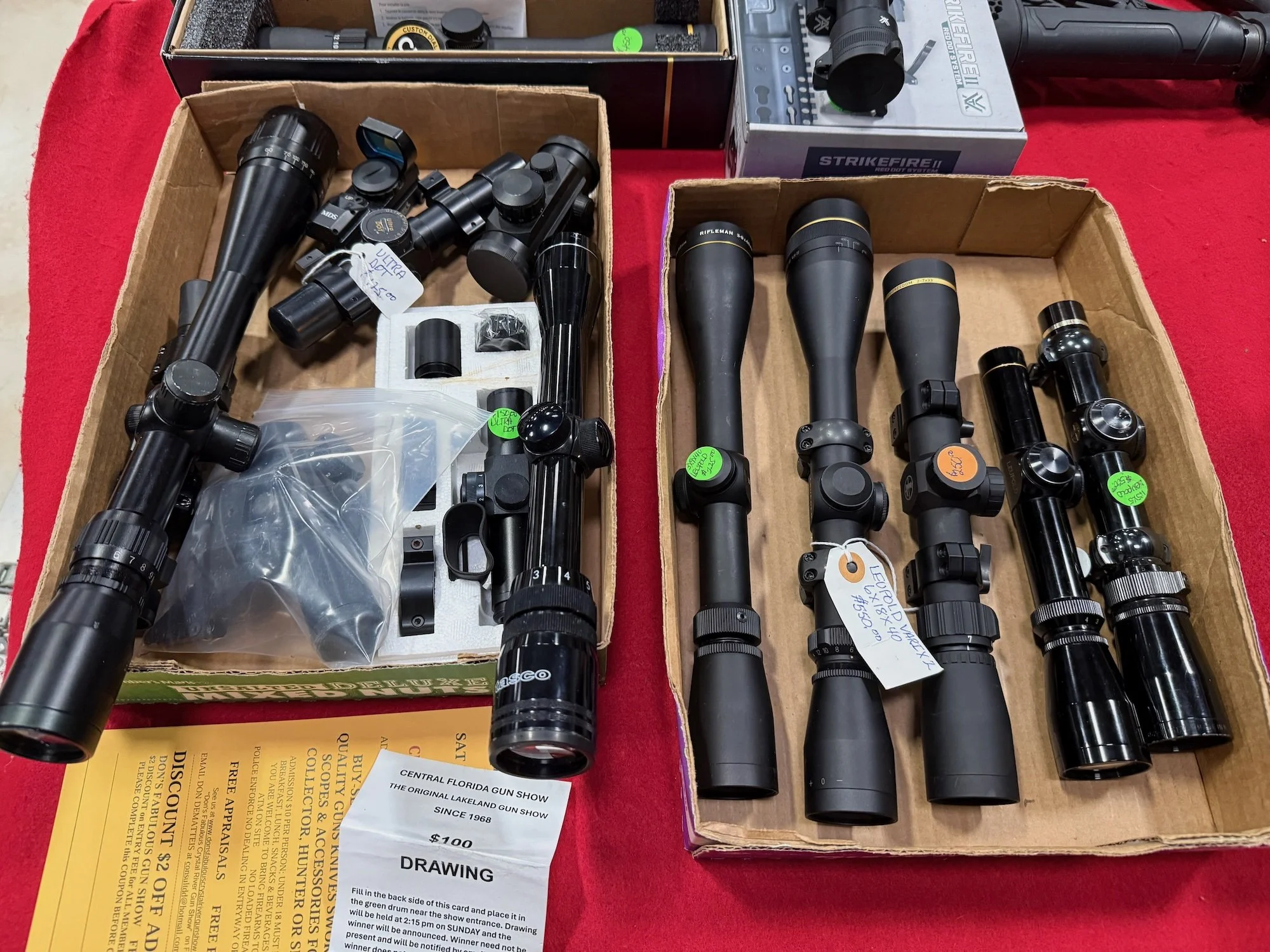 A collection of rifle scopes and accessories displayed on a red tablecloth, with handwritten and printed price tags attached.