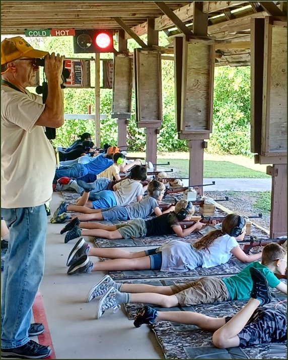 Children lying prone on the ground at a shooting range, aiming rifles at targets, with an instructor or coach observing. An outdoor setting with a wooden shooting shelter and a light signal with 'Cold' and 'Hot' indicators.