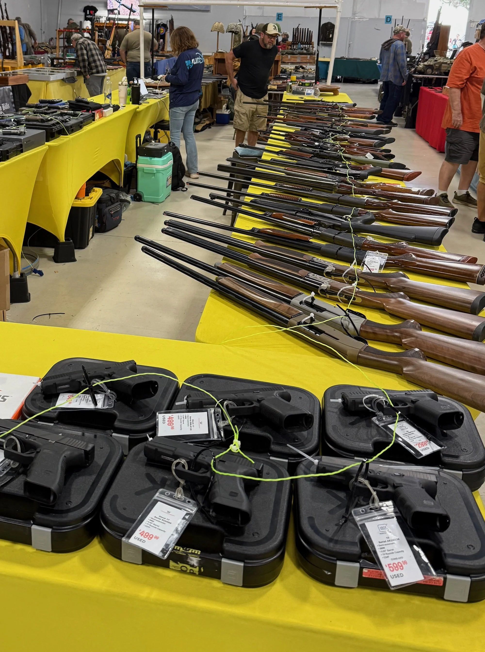 A table with multiple handguns in black cases and long rifles on display at a gun show. The background shows people browsing other tables with various firearms and accessories.
