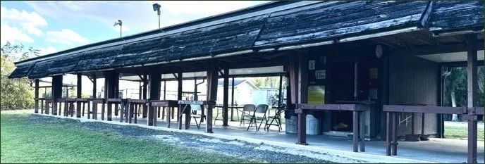 Long outdoor wooden pavilion with tables and chairs under a metal roof, surrounded by grassy area and trees.