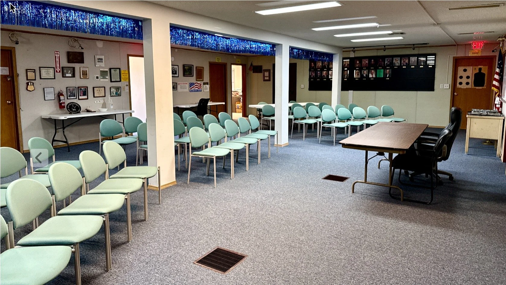 Empty meeting room with rows of light green chairs, a brown table with black chairs, and a wall of photographs and certificates.
