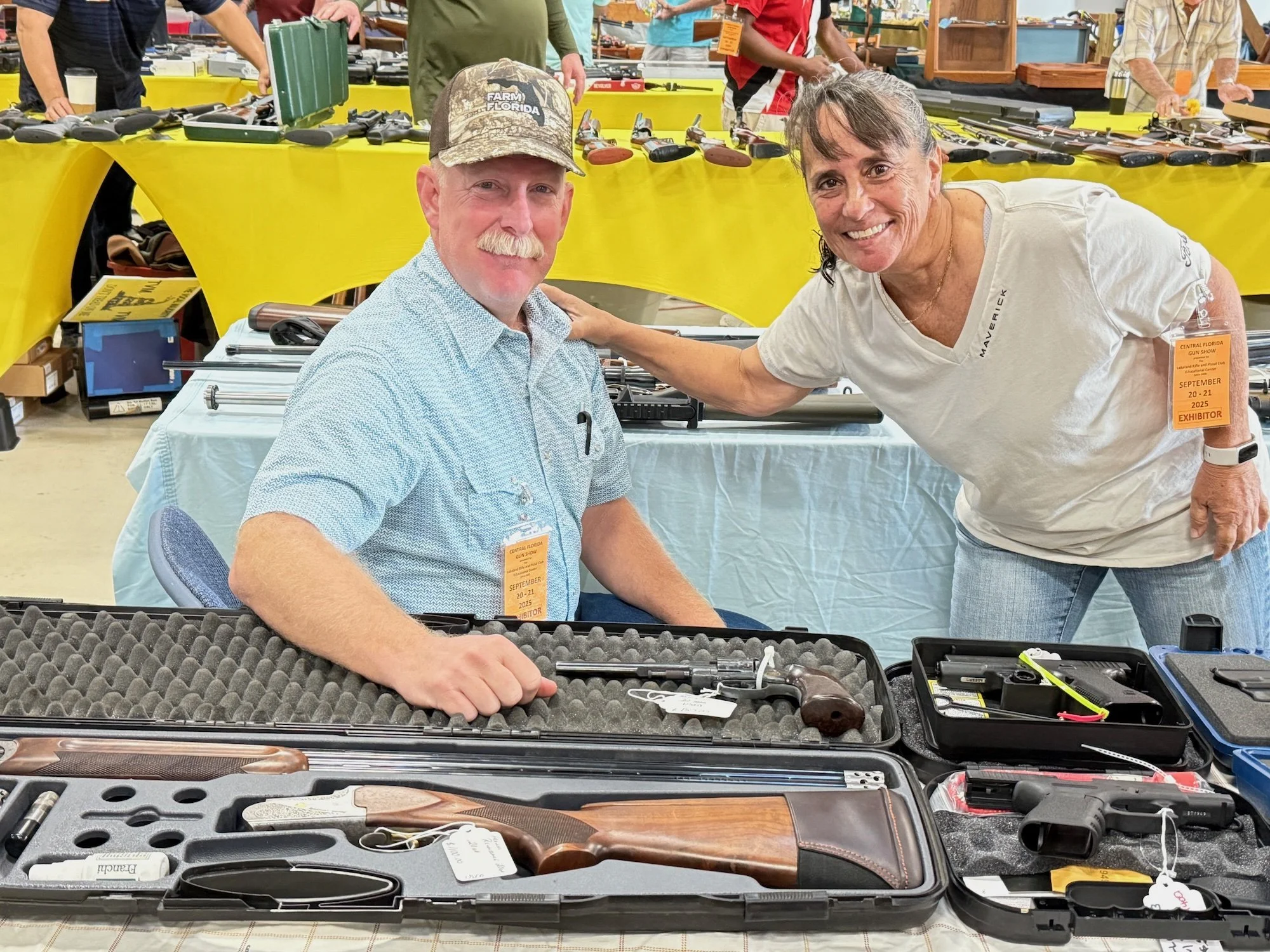 A man and woman at a gun show with guns on display at their booth. The man, seated, wears a blue shirt and cap, and the woman, standing, wears a white shirt. They are smiling, and the woman has her arm around the man. The booth has a yellow tablecloth and various firearms in cases.