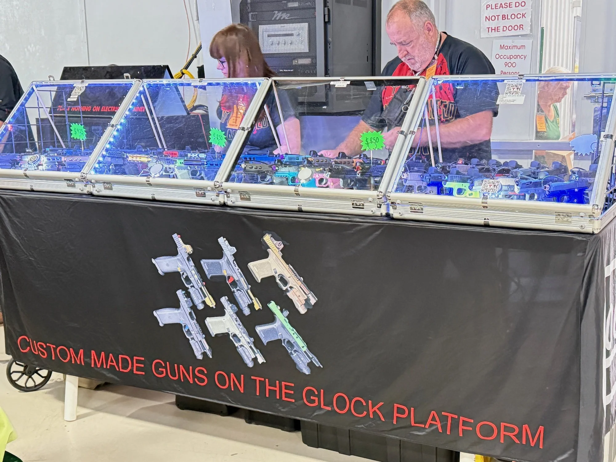 A booth displaying custom-made guns on a black tablecloth with a sign that reads 'Custom Made Guns on the Glock Platform.' A woman and a man vendor are standing behind the glass display case.
