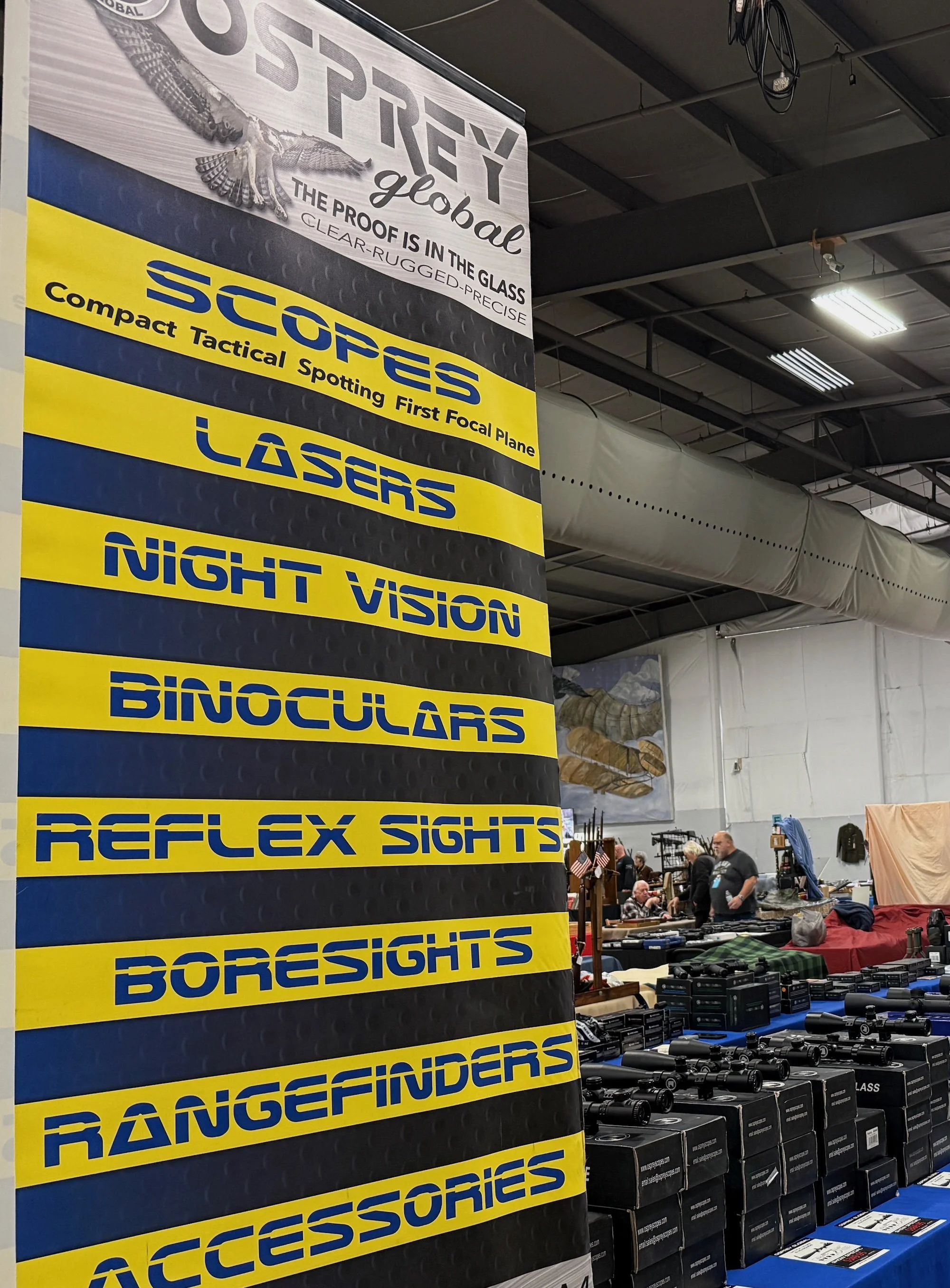 A large display banner lists various hunting and shooting accessories including scopes, lasers, night vision, binoculars, reflex sights, boresights, rangefinders, and accessories, at a gun show.