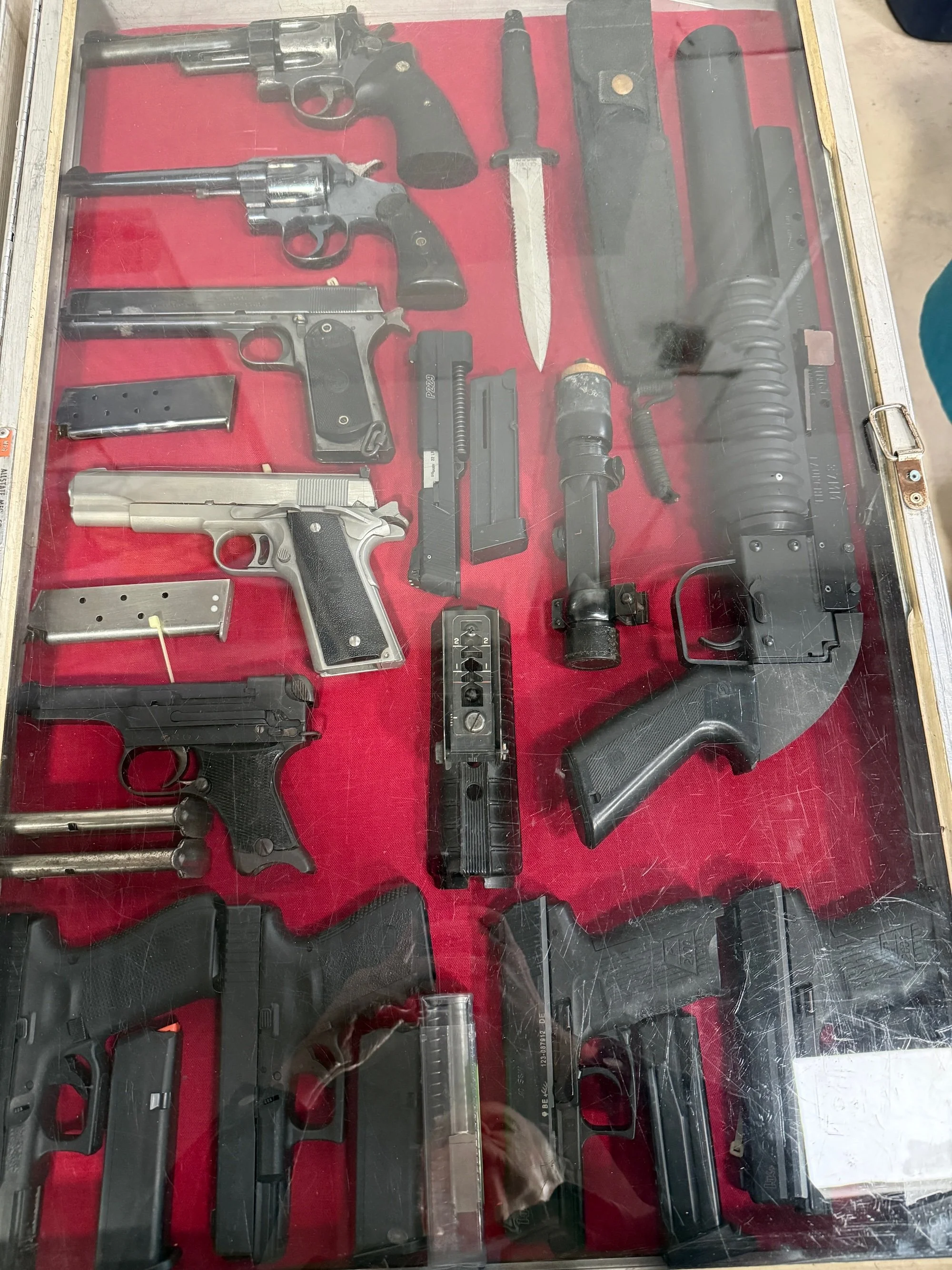 Display of various firearms including revolvers, semi-automatic pistols, a rifle, a knife, and magazines, arranged on a red background inside a glass case.