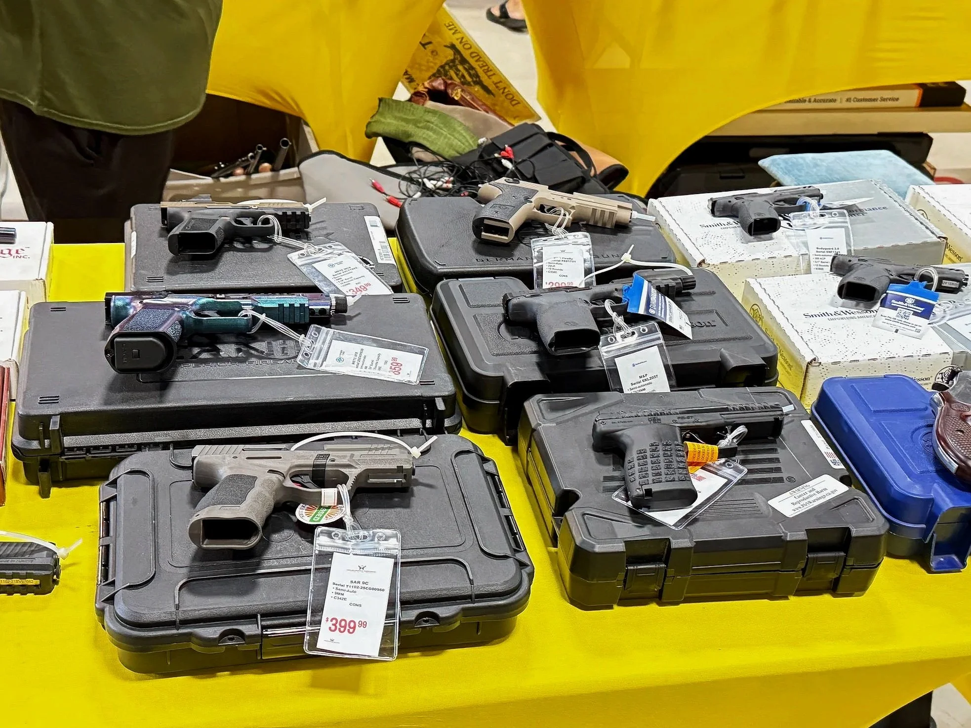 Table displaying multiple handguns, some inside black carrying cases, with price tags attached. The background includes yellow tablecloths and various other firearm accessories.