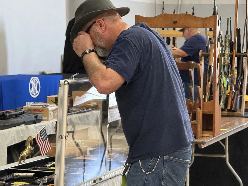 Man leaning forward, touching his glasses to look closely at at something in a display case. Small US flag is visible. Rifles in display stands in background.
