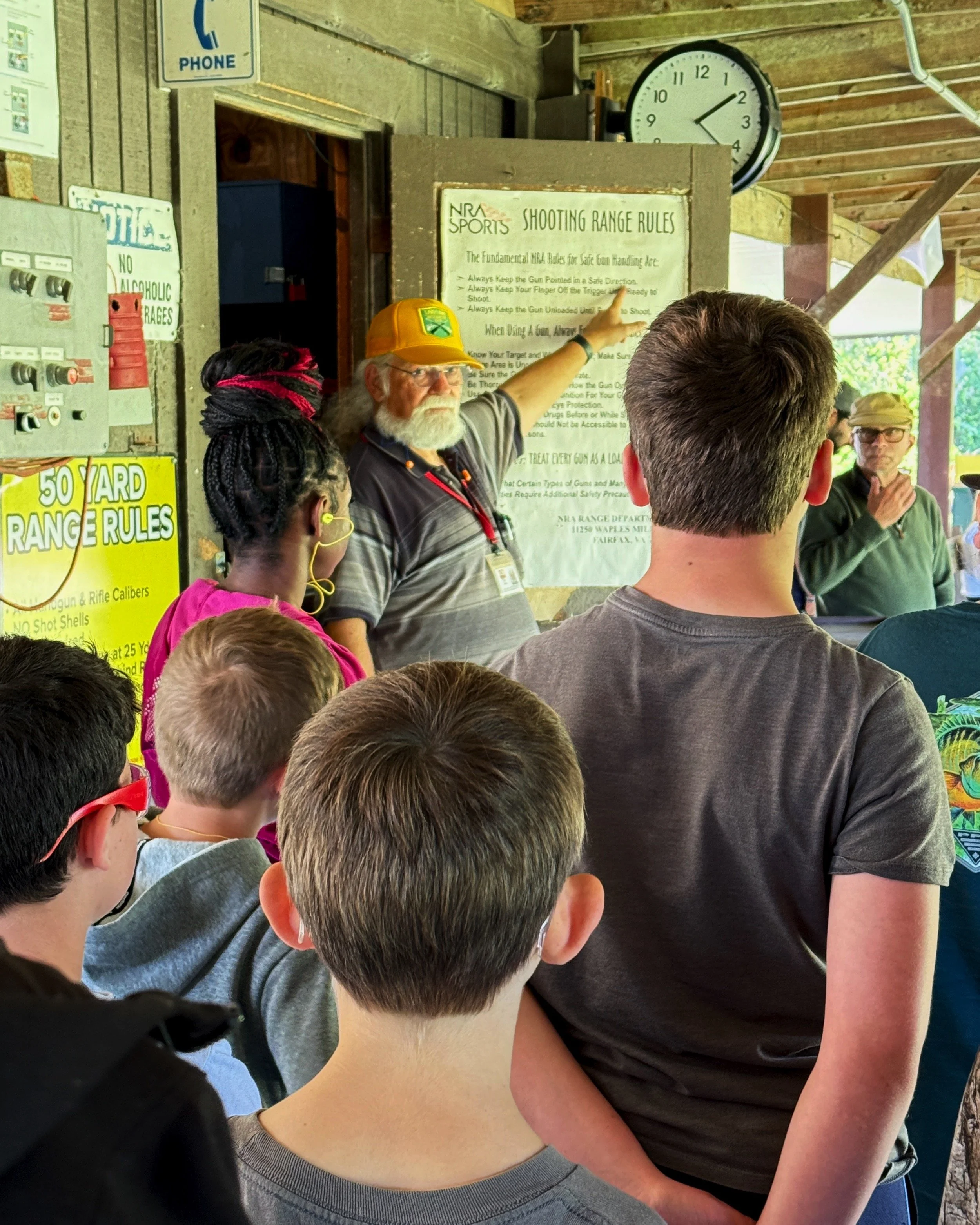 An instructor with a yellow Lakeland Rifle & Pistol Club hat is explaining range safety rules to children at an outdoor shooting range. The instructor is pointing at a sign with safety rules, and a clock is visible in the background.