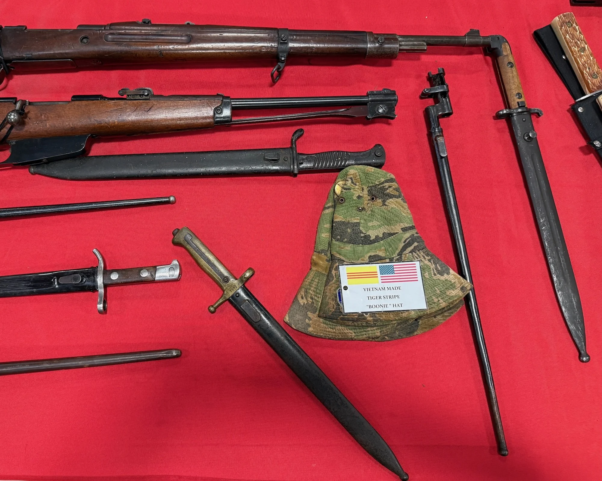 Collection of vintage military weapons and a camouflage hat with a label indicating Vietnam-made tiger stripe boonie hat, displayed on a red surface.