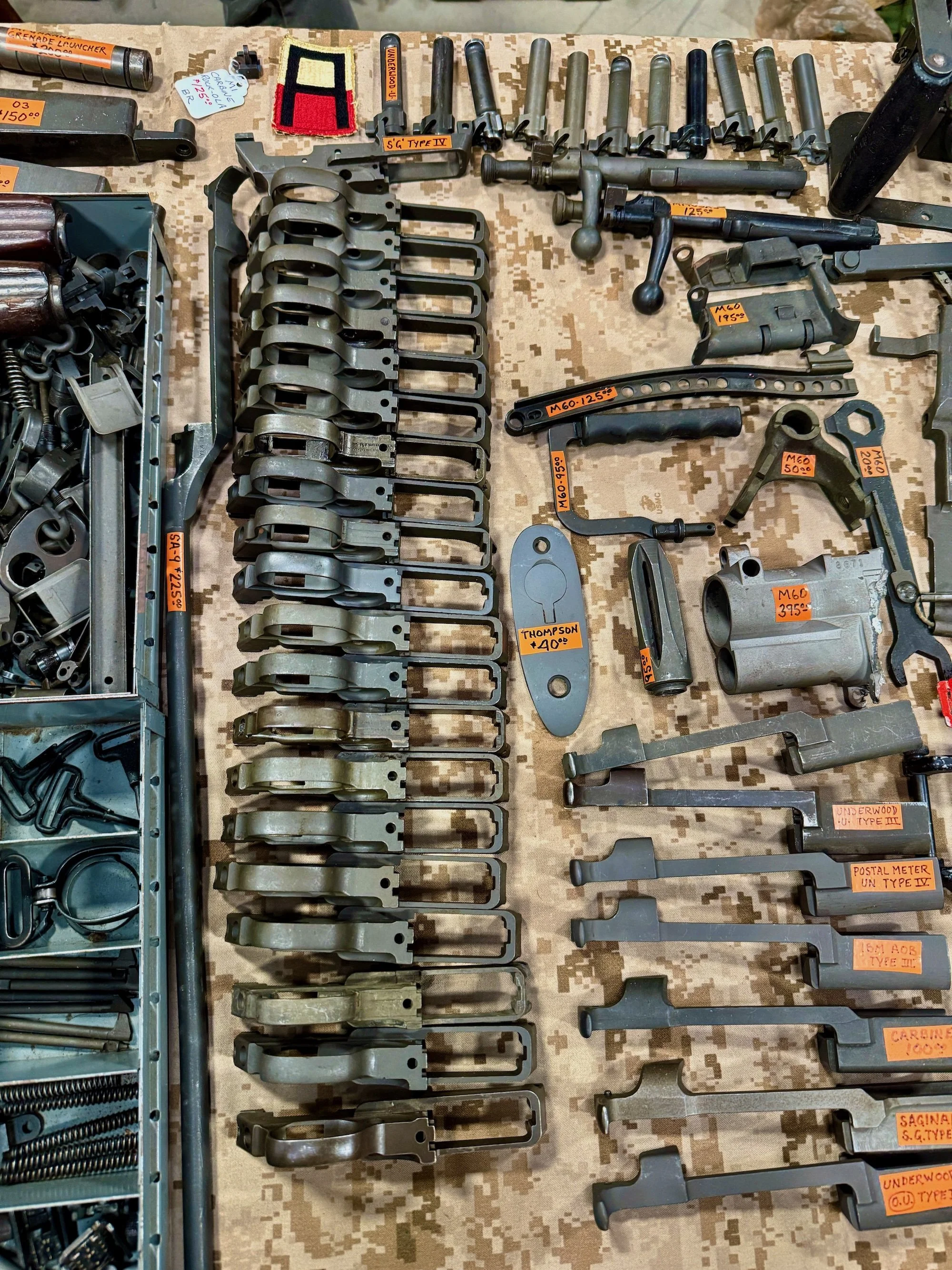 Collection of military firearm parts and accessories, including bolt carriers, bolts, and tools, laid out on a camouflage cloth.