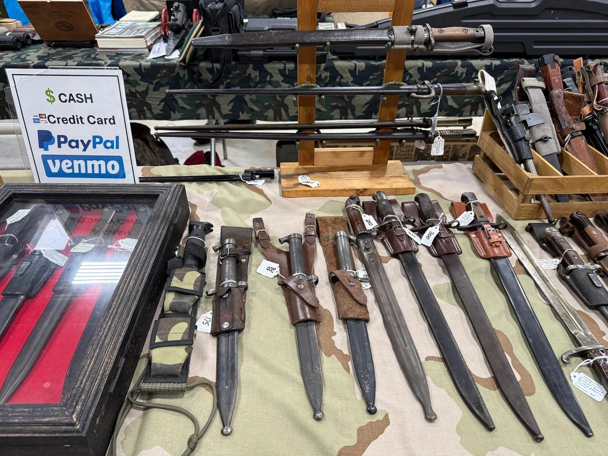 Display of vintage bayonets and knives on a camouflage tablecloth at a gun show, with signs indicating cash, credit card, PayPal, and Venmo payment options.