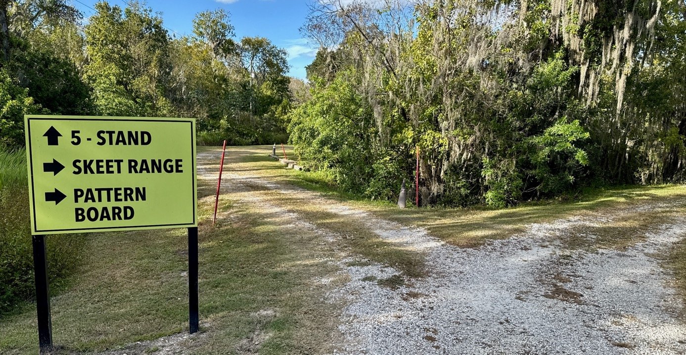 A gravel path leading through a wooded area with a sign on the left indicating directions for a skeet range, pattern board, and stand.