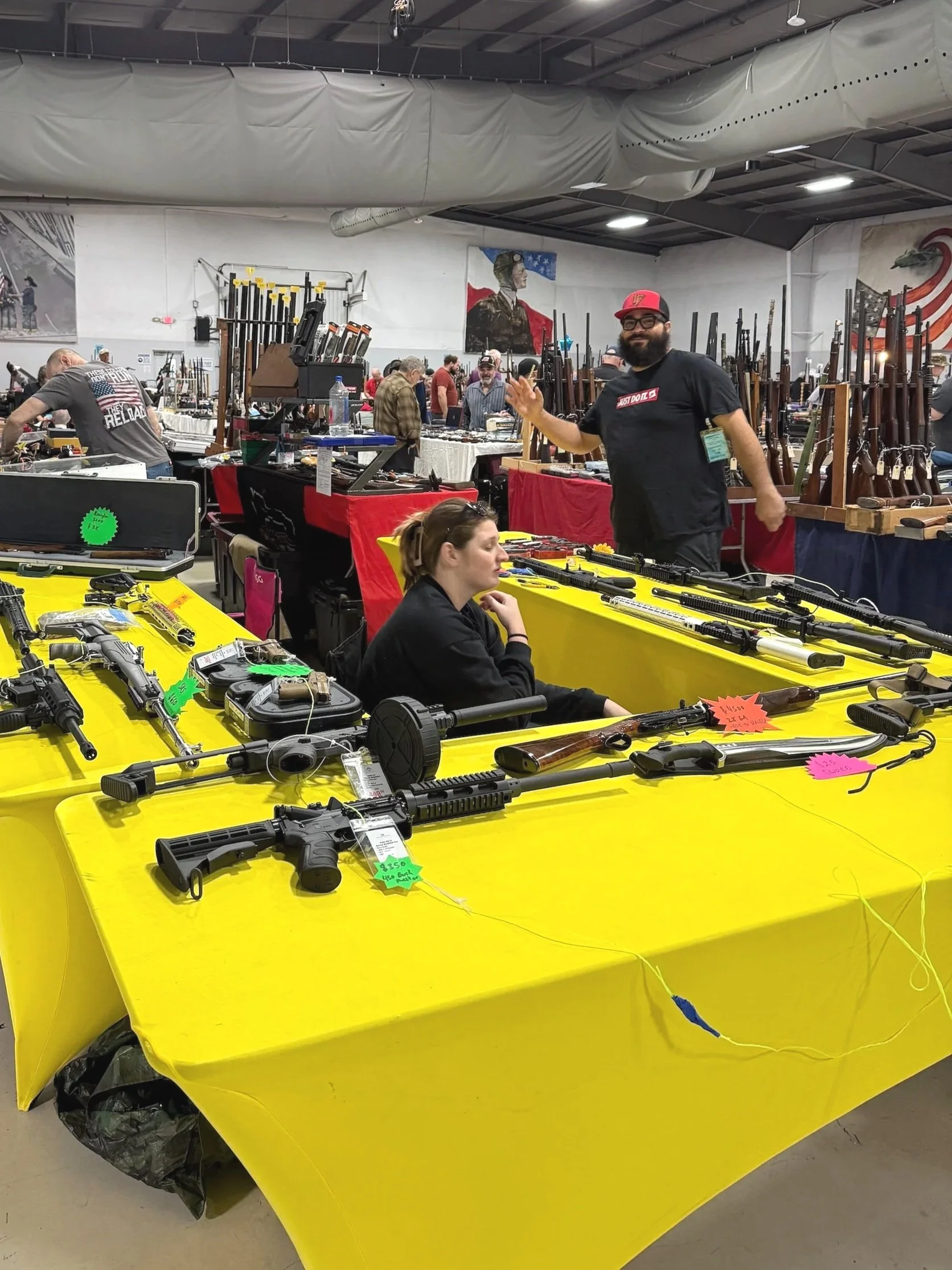 At a gun show, a man and a woman are manning a booth at a gun show with various firearms on display, with other vendors and visitors in the background.