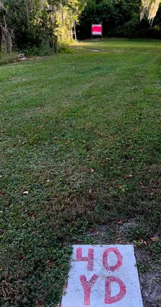 A grassy outdoor area with trees in the background at a gun club. Pattern board in the distance with 40 yd distance marker.