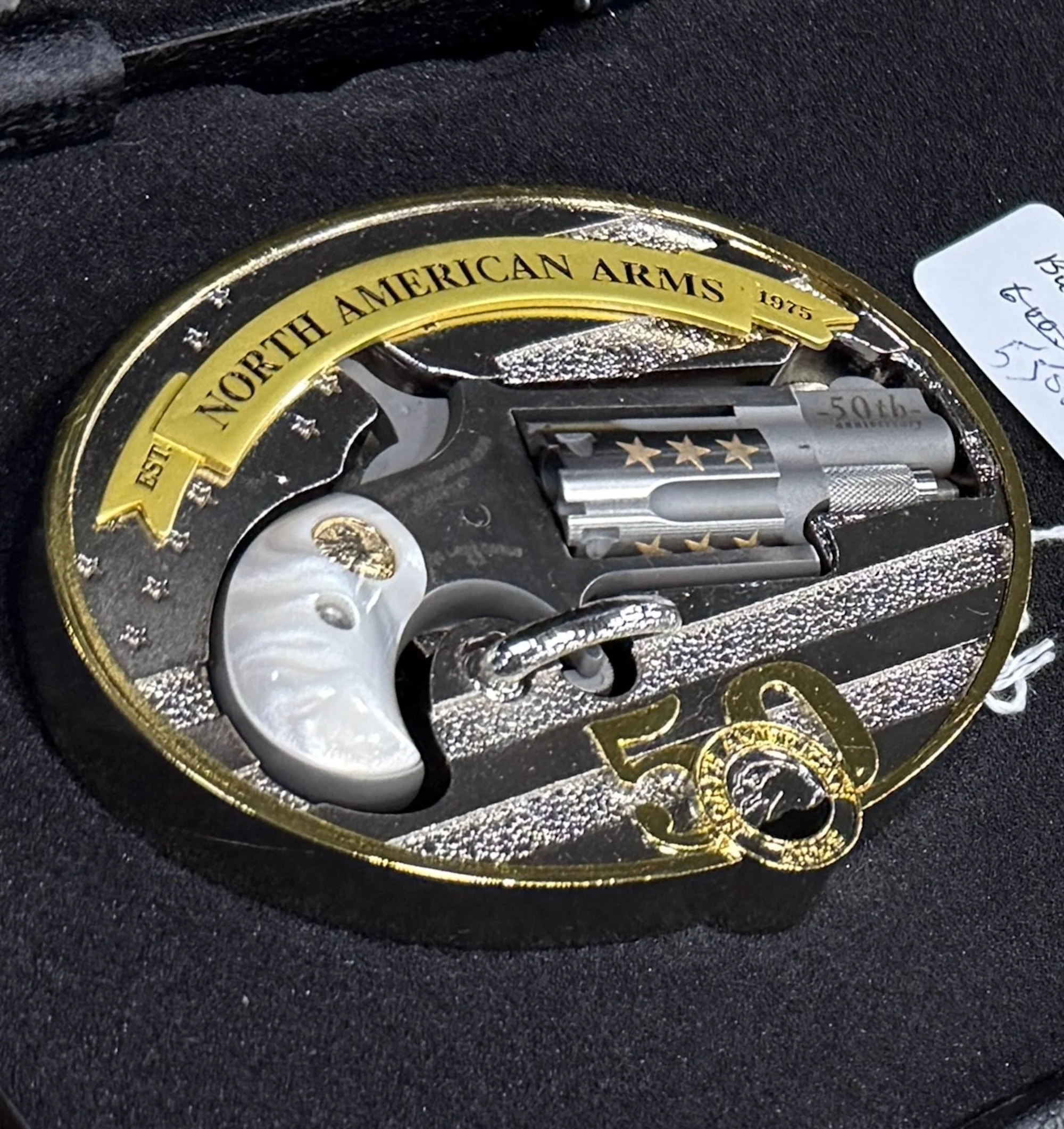 Decorative commemorative display with a revolver-shaped piece, a white pearl-like object, and a small engraved card, celebrating the 50th anniversary of the North American Arms company, established in 1975, featuring a gold and black oval frame with stars and a banner.