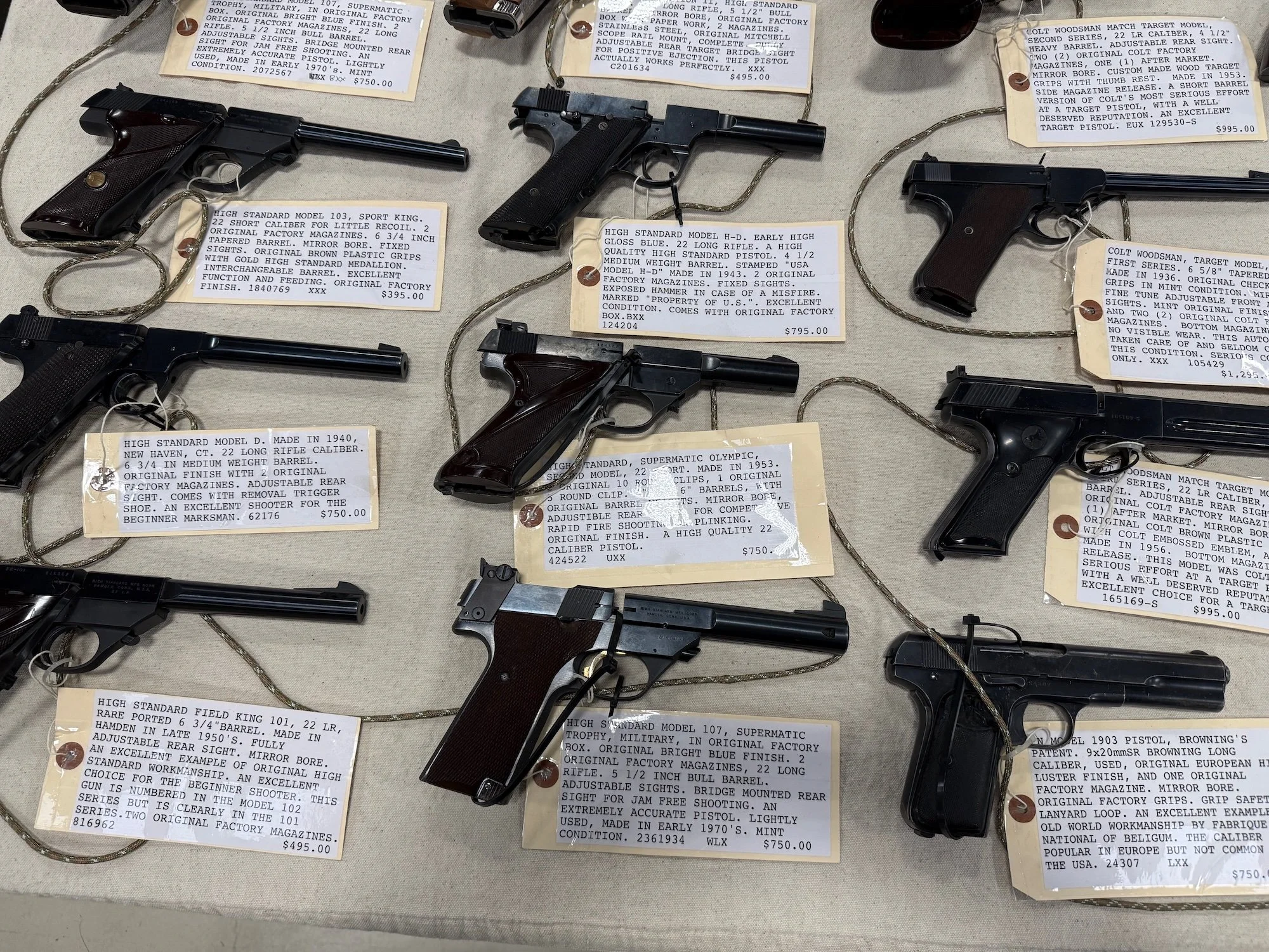 Table displaying various vintage handguns, each with a tag detailing specifications, origin, and price. The guns include models from manufacturers such as Colt and High Standard, with descriptions highlighting features, history, and condition.
