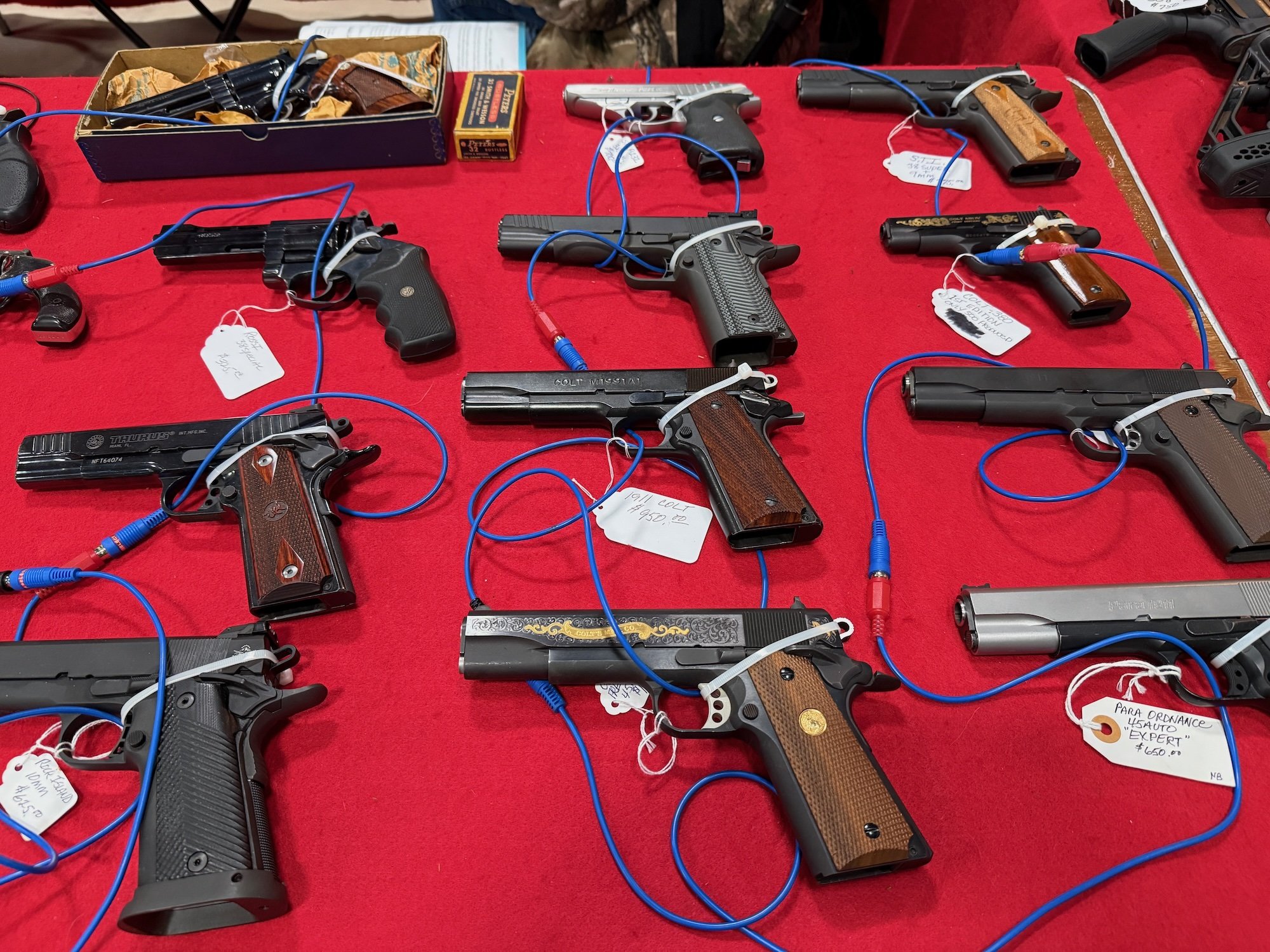 Multiple handguns displayed on a red tablecloth, connected with blue and white security tags, with some tags showing prices and descriptions.