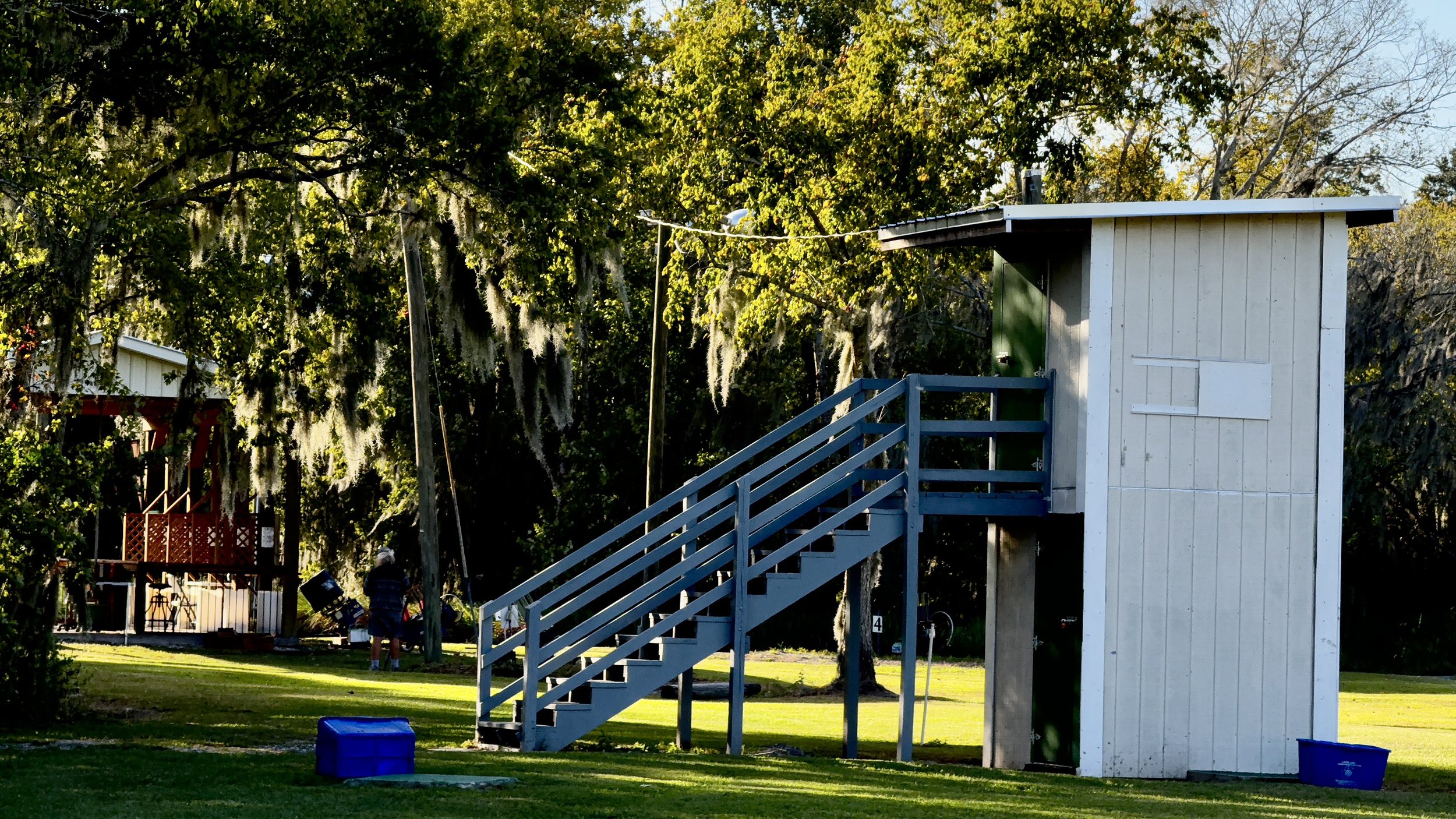 A small white wooden structure with a blue staircase, elevated on stilts, surrounded by lush green trees with Spanish moss at a gun club. Five-stand building in the background.