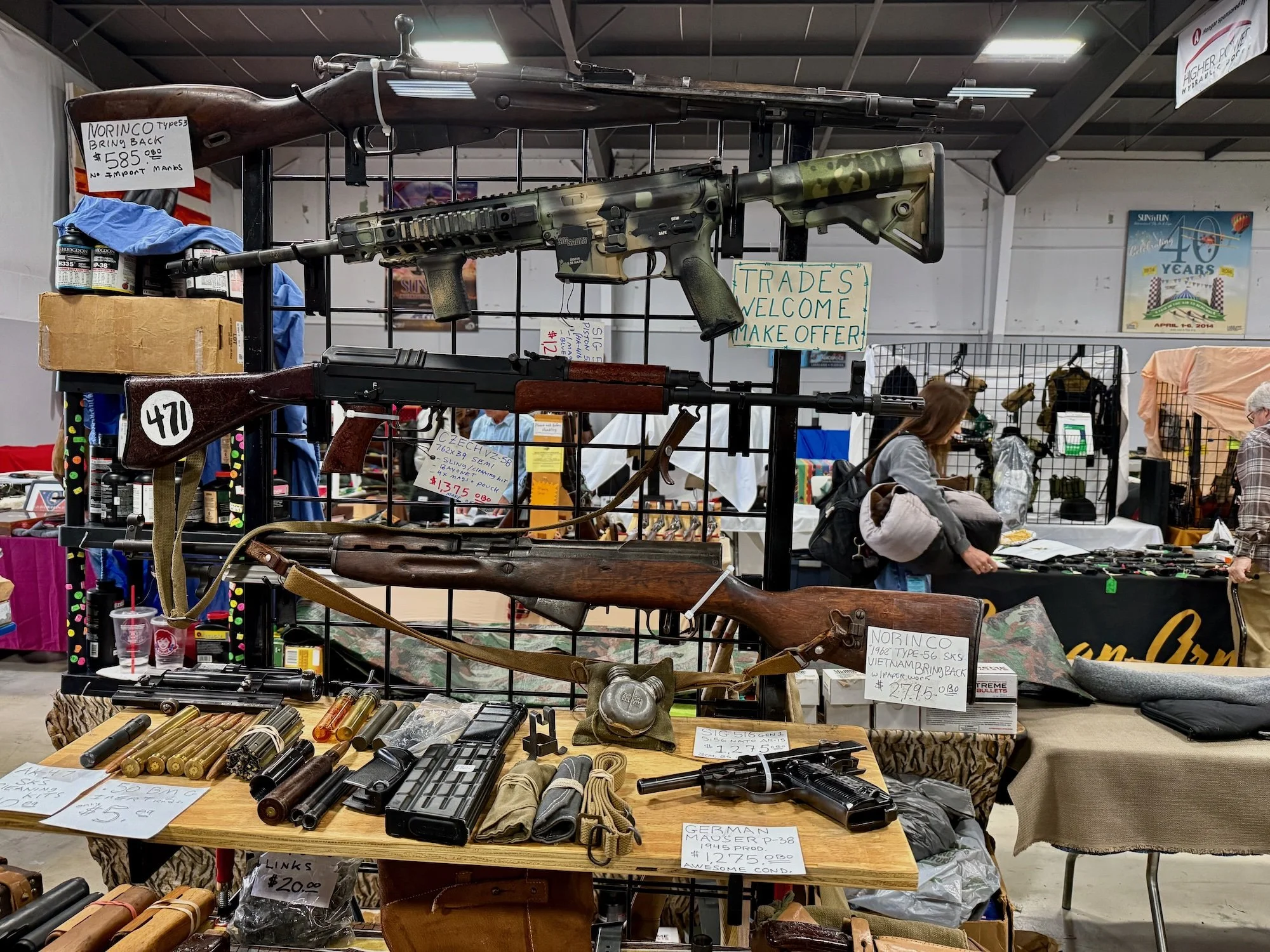 Display of vintage firearms including rifles, pistols, and ammunition at a gun show, with signs indicating prices and descriptions, and people browsing in the background.