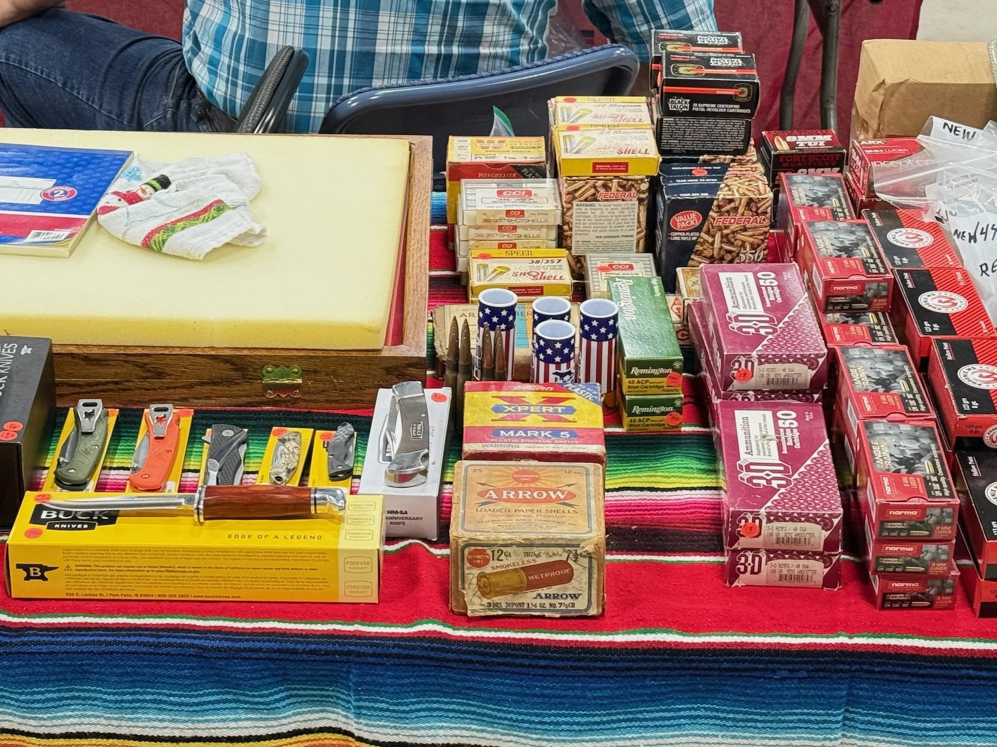 A table displaying various ammunition boxes, knives, and small items arranged on a colorful striped tablecloth. Items include shotshells, bullets, and small utility knives.