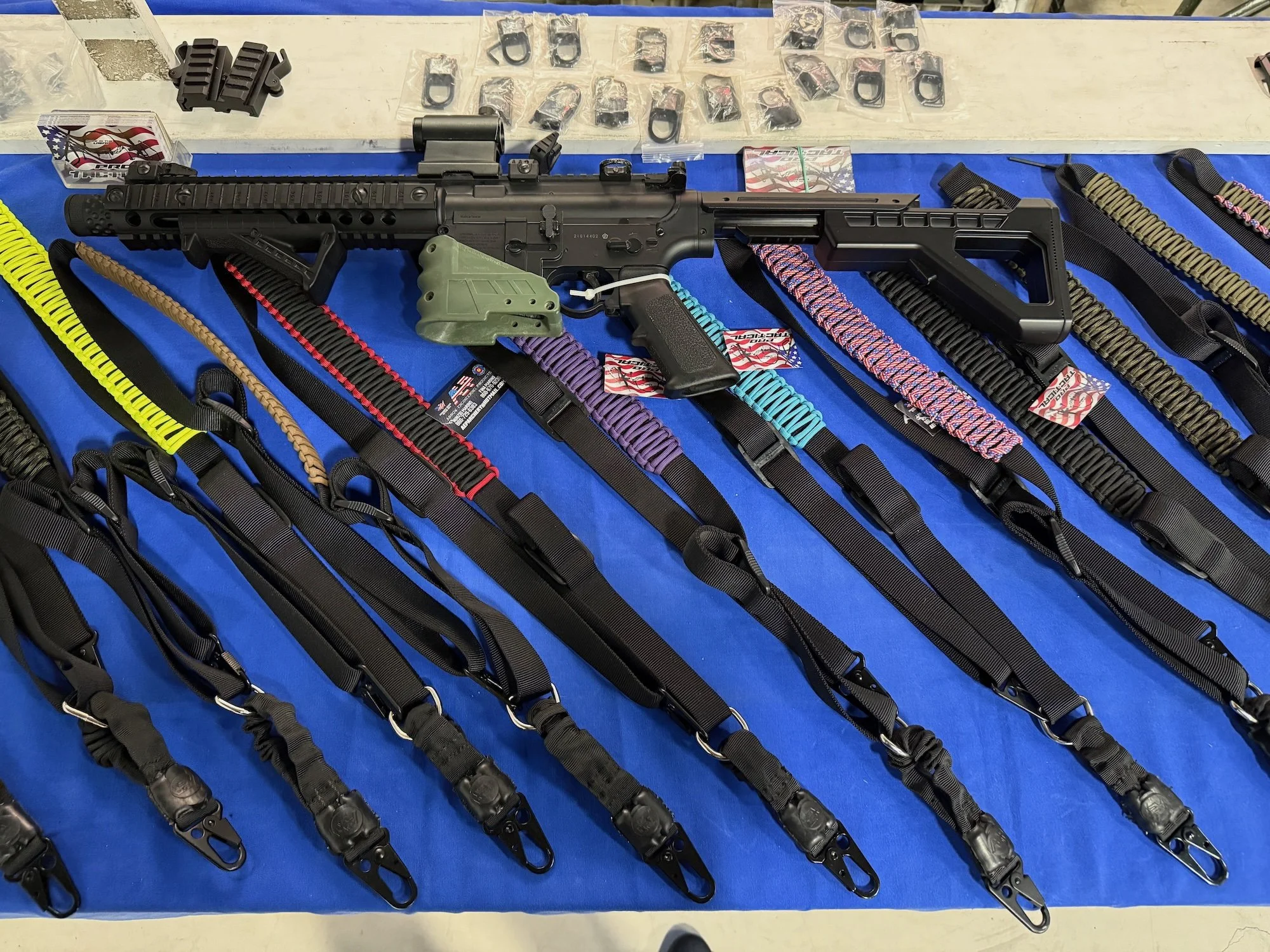 A display of a black semi-automatic rifle with a green grip, laying on a blue cloth, with various colorful strap slings arranged in front of it, and small plastic bags containing firearm accessories in the background.