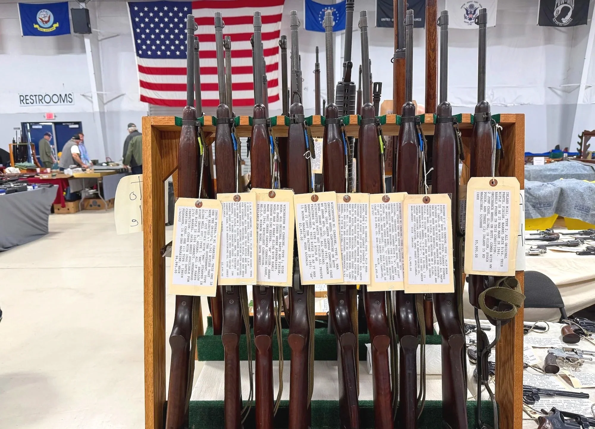 A display of antique rifles with price tags on a wooden stand at a gun show, with American flags and other booths in the background.