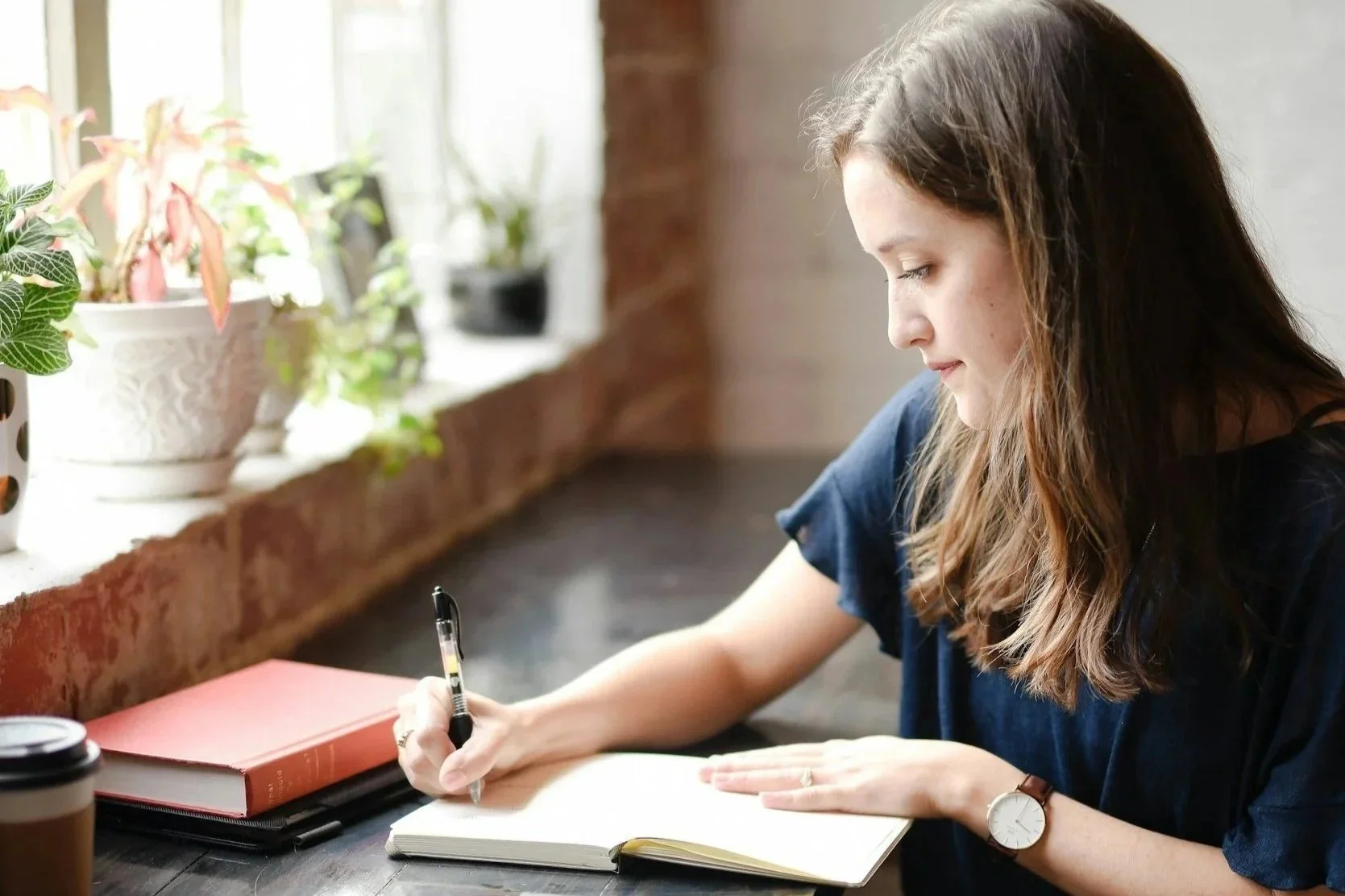 Young woman with long brown hair writing in a notebook at a desk with plants on the window sill.