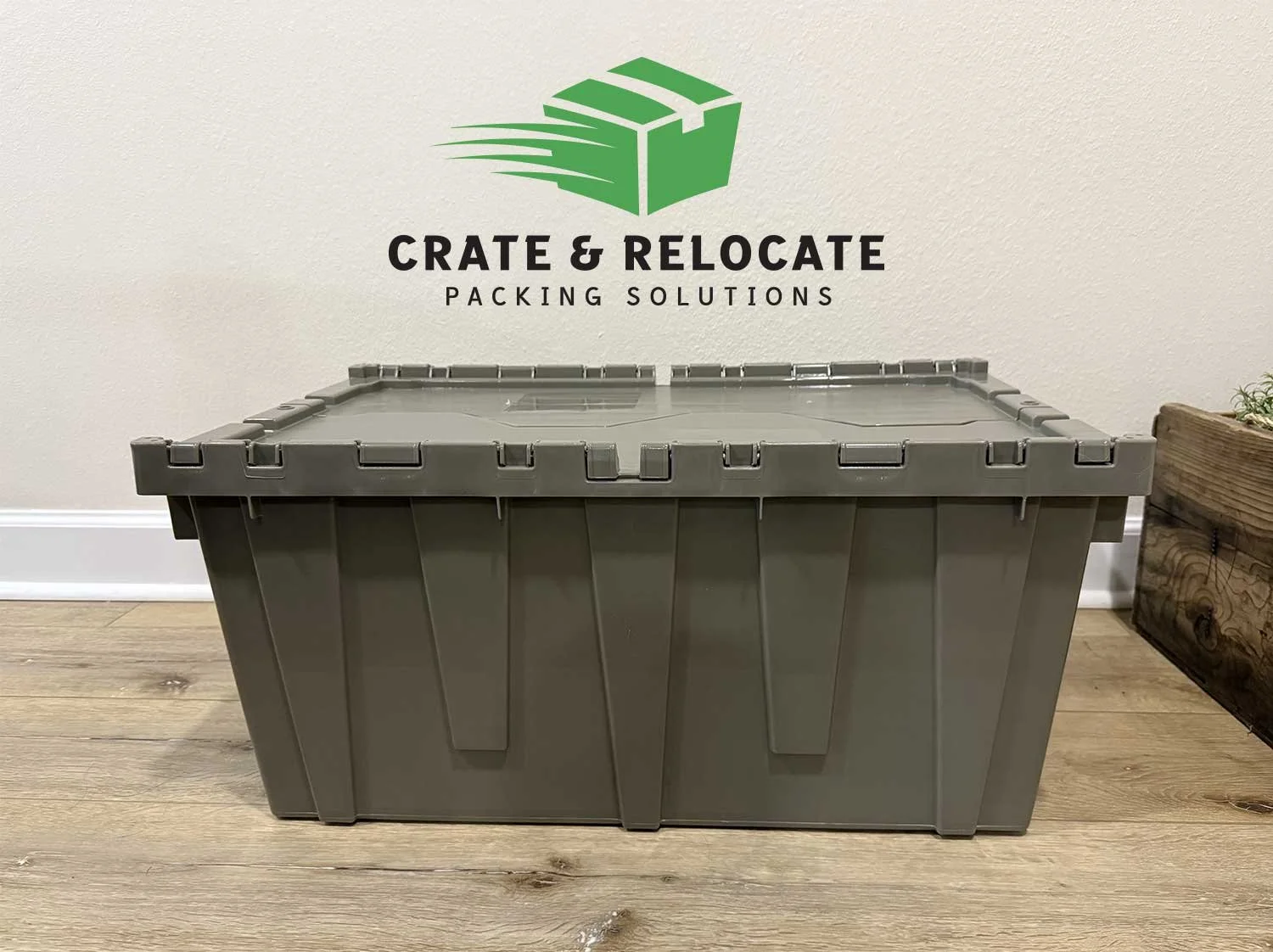A large gray plastic storage container in front of a wall with a company logo reading 'Crate & Relocate Packing Solutions.' The container is on a wooden floor, and there is a wooden planter box with some plants on the right side.