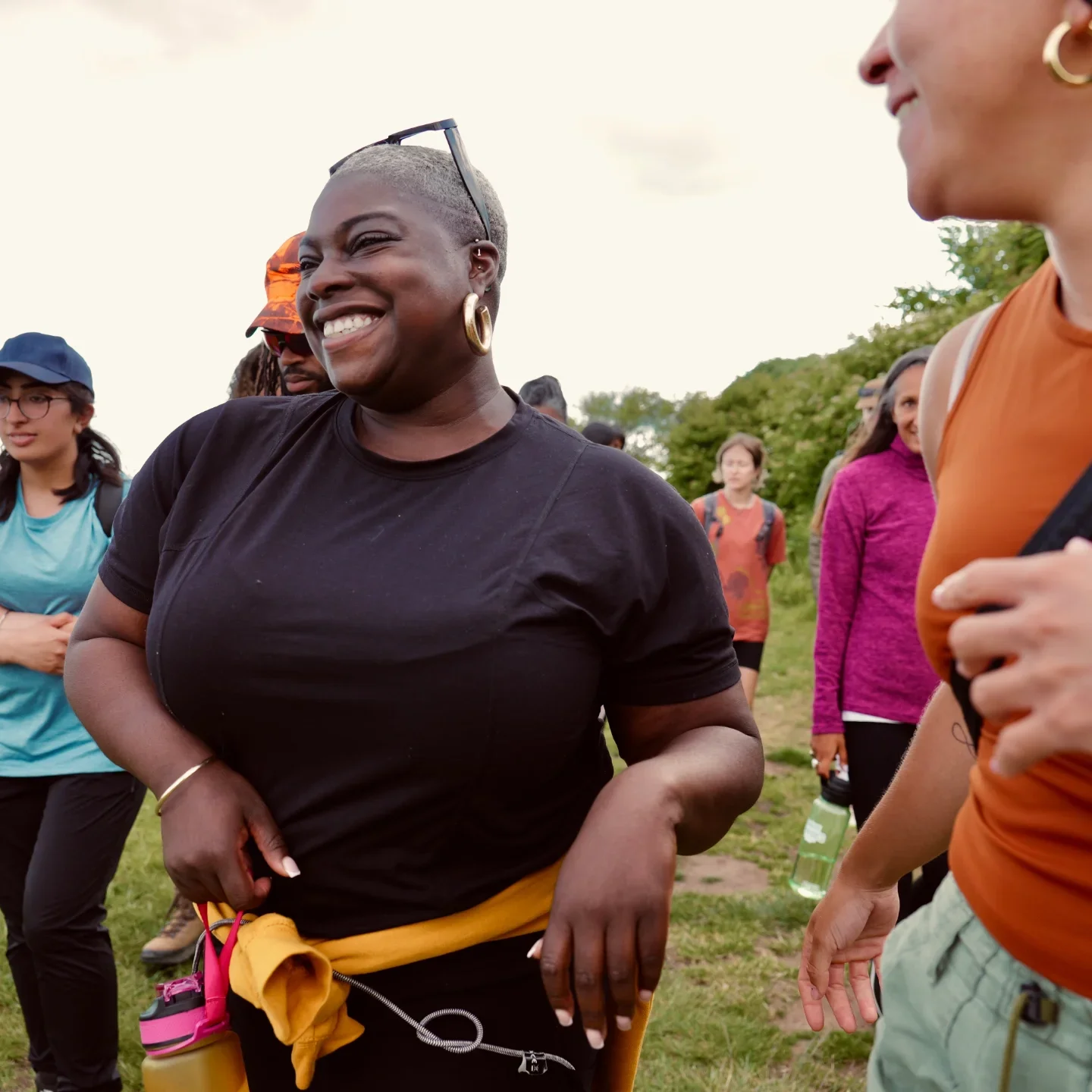 A group of diverse women standing outdoors, smiling and chatting in a park setting during daytime.