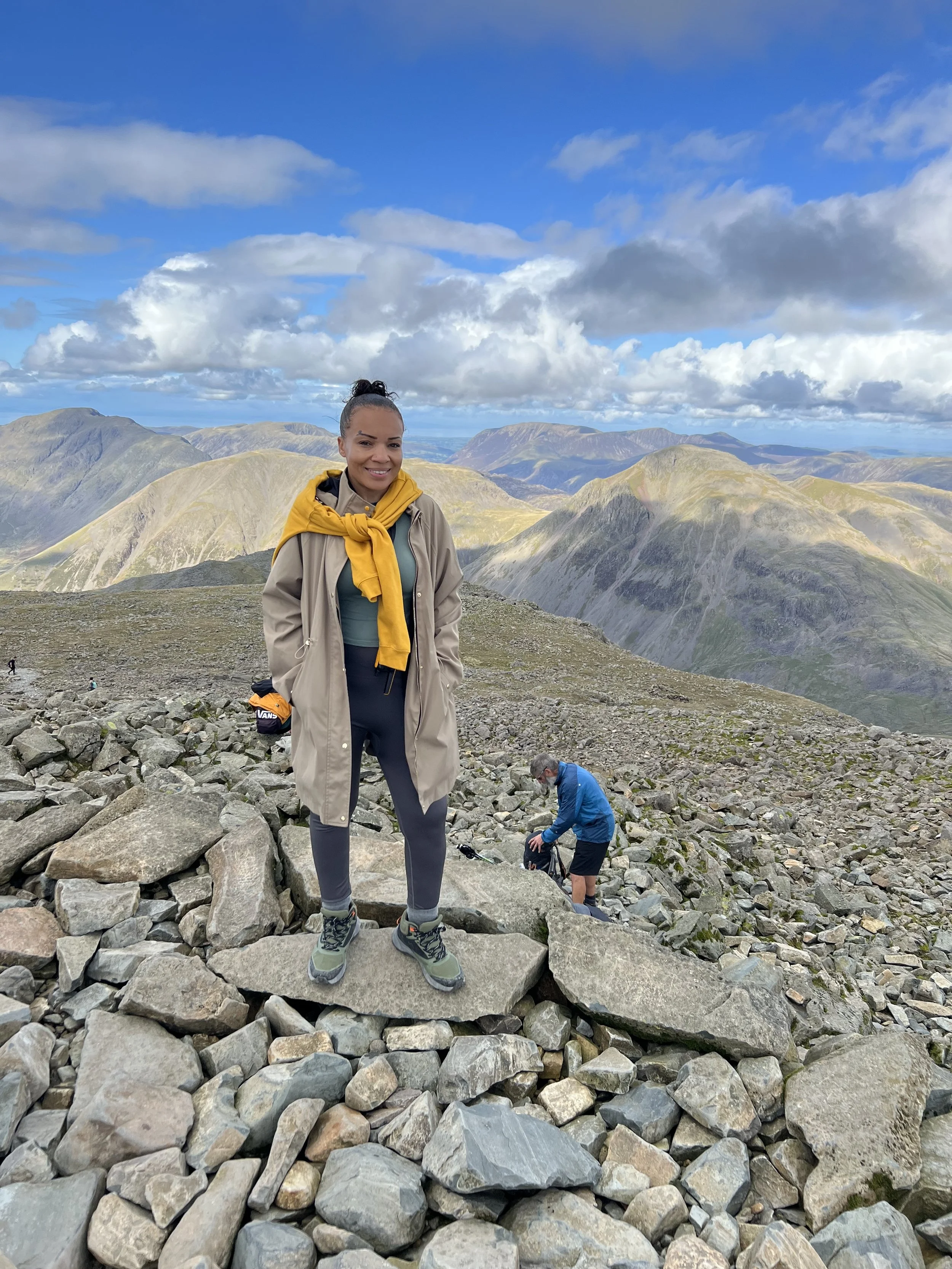 A woman standing on rocky terrain during a hike in a mountainous area, with mountains and a partly cloudy sky in the background.