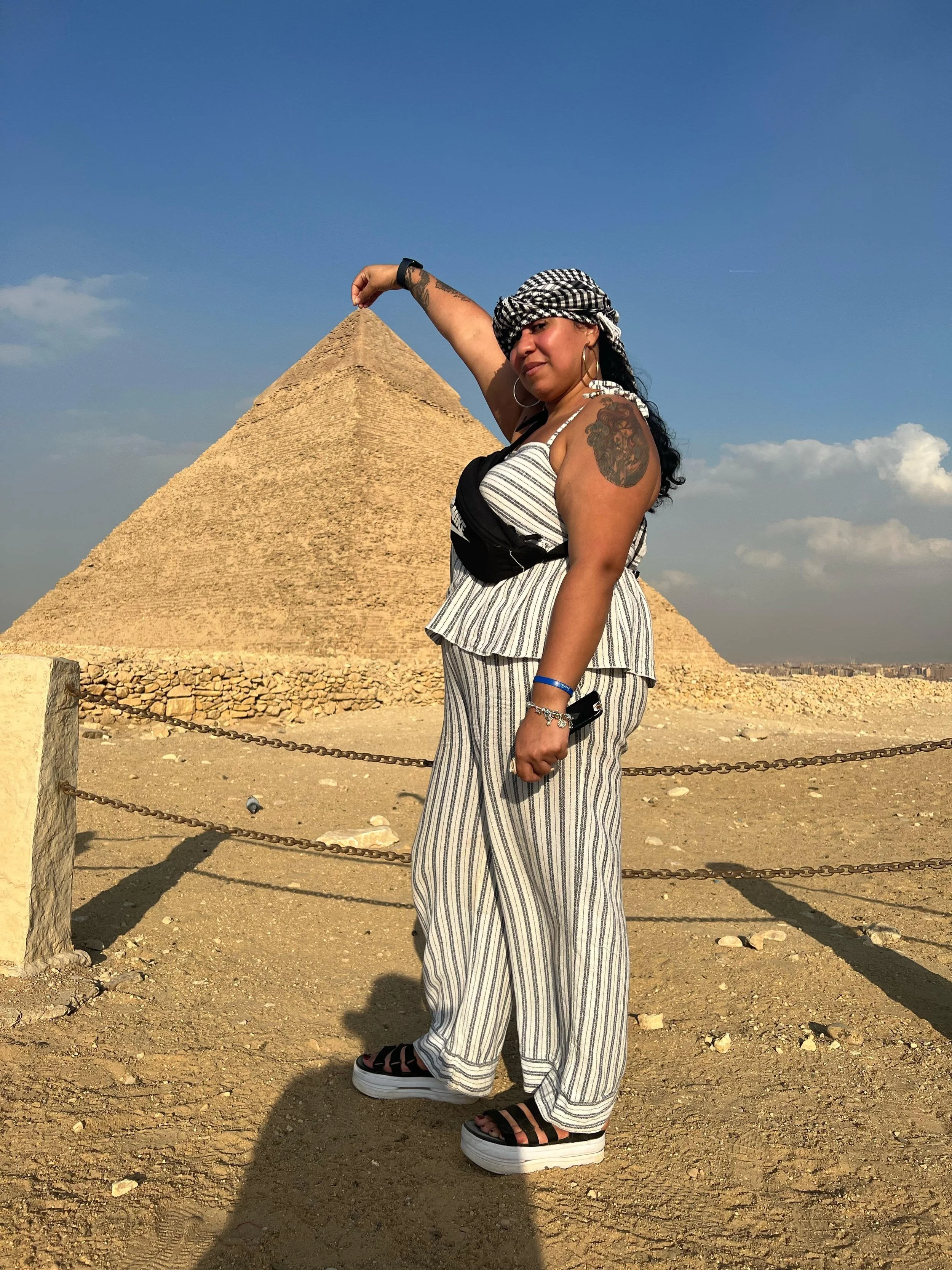 Woman posing in front of a pyramid, wearing striped outfit, sandals, and a keffiyeh headscarf.