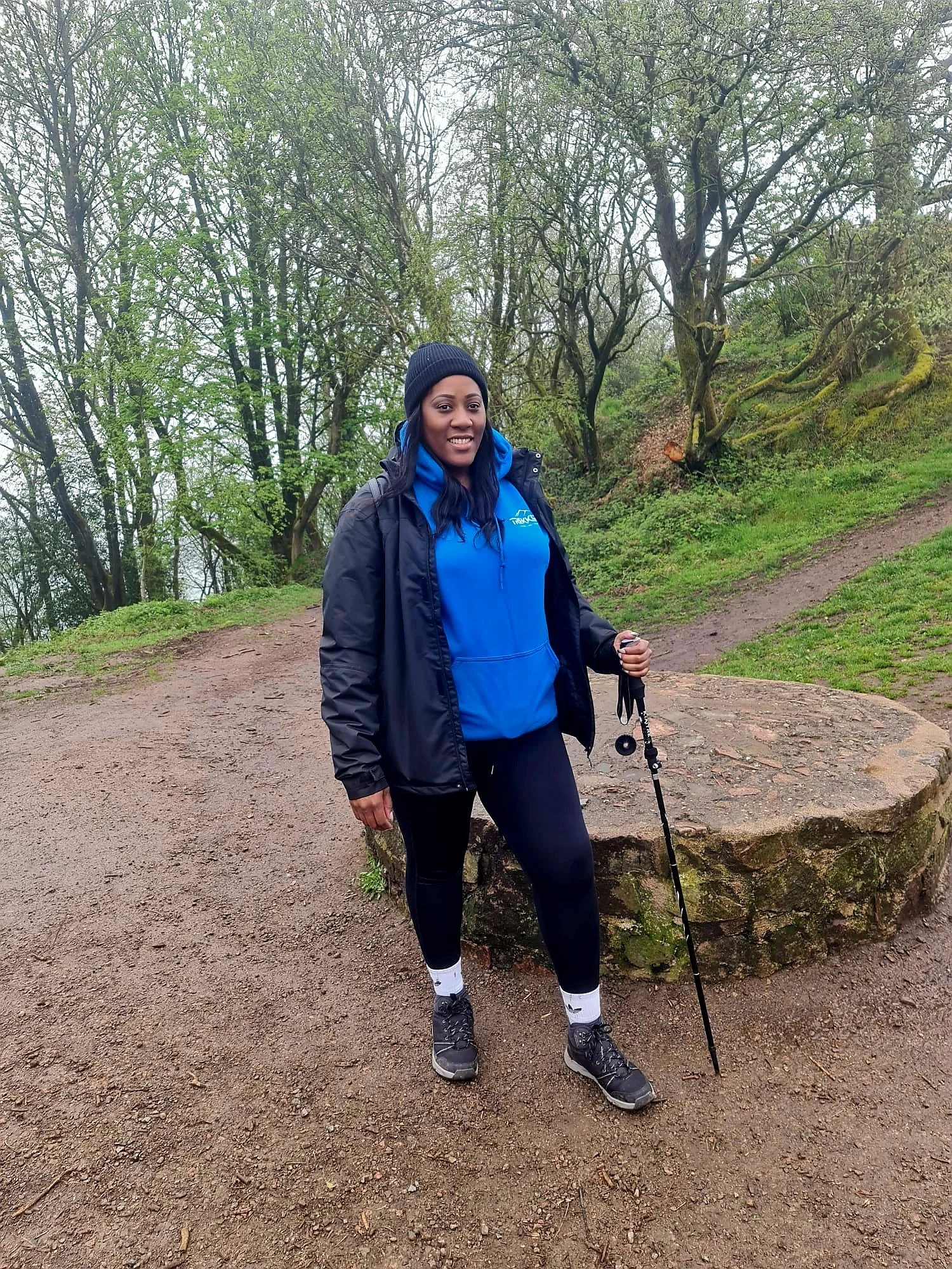 A woman dressed in hiking gear standing on a dirt trail in a wooded area with trees and green foliage.