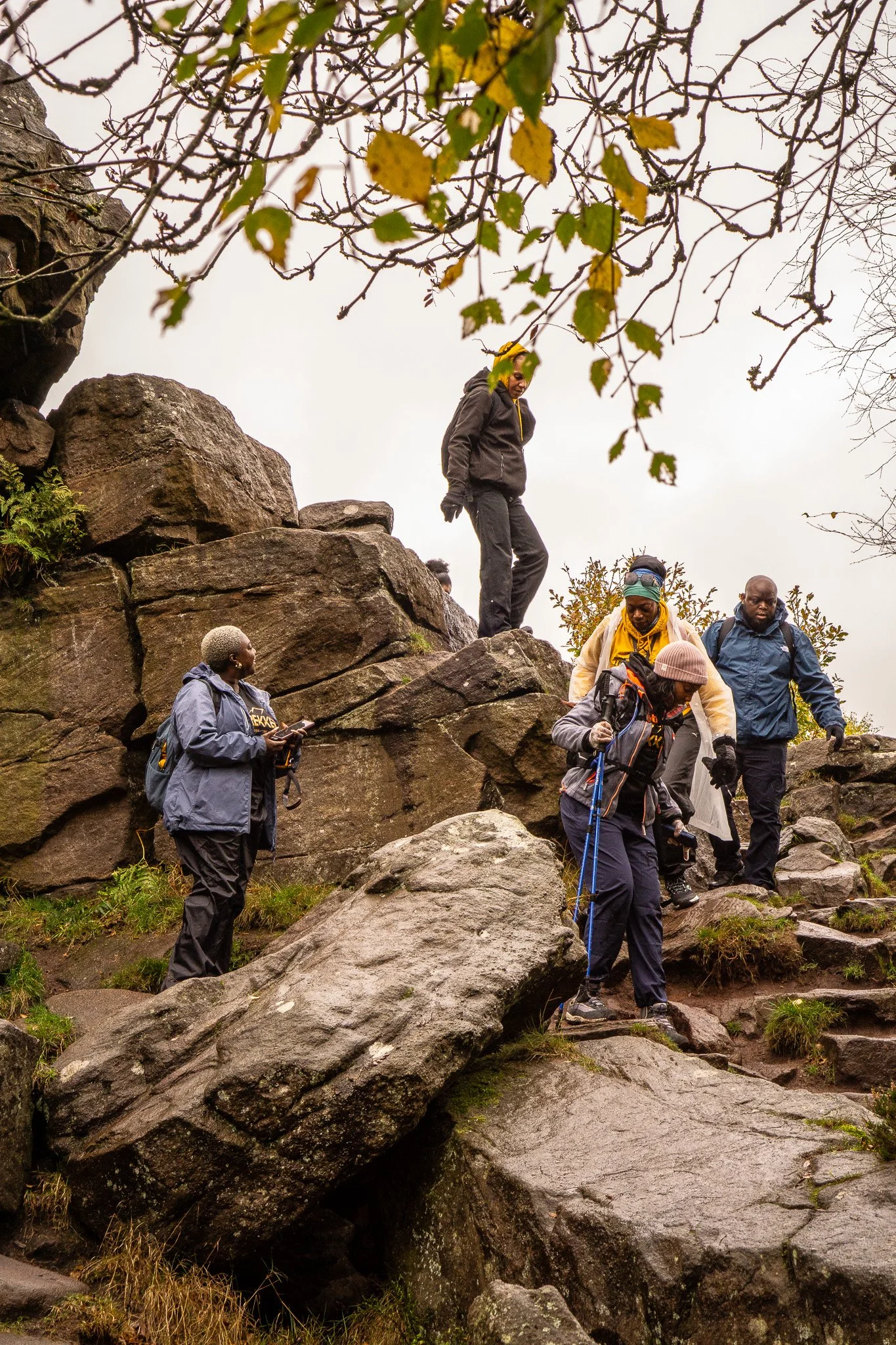 Group of hikers climbing over large rocks on a mountain trail, dressed in outdoor gear and carrying backpacks during cloudy weather.