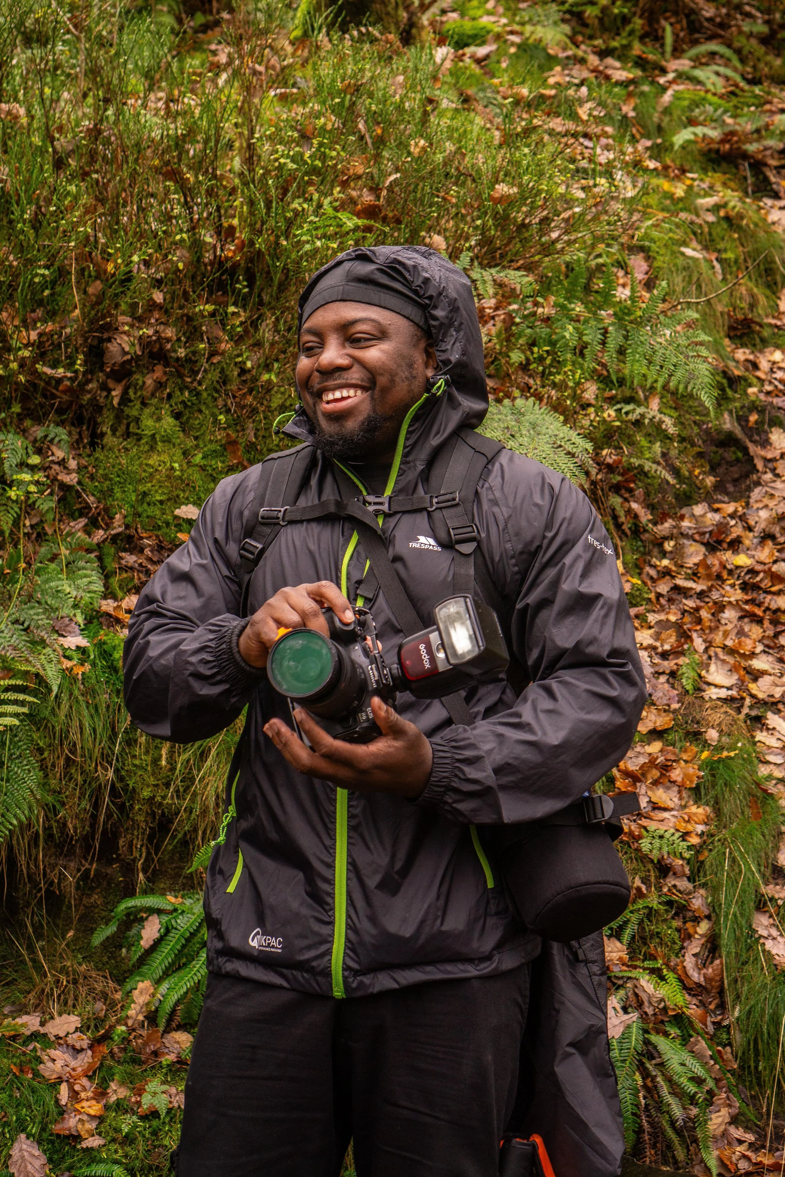 A man dressed in black outdoor gear holding a camera, smiling, standing in a forest with ferns and fallen leaves.