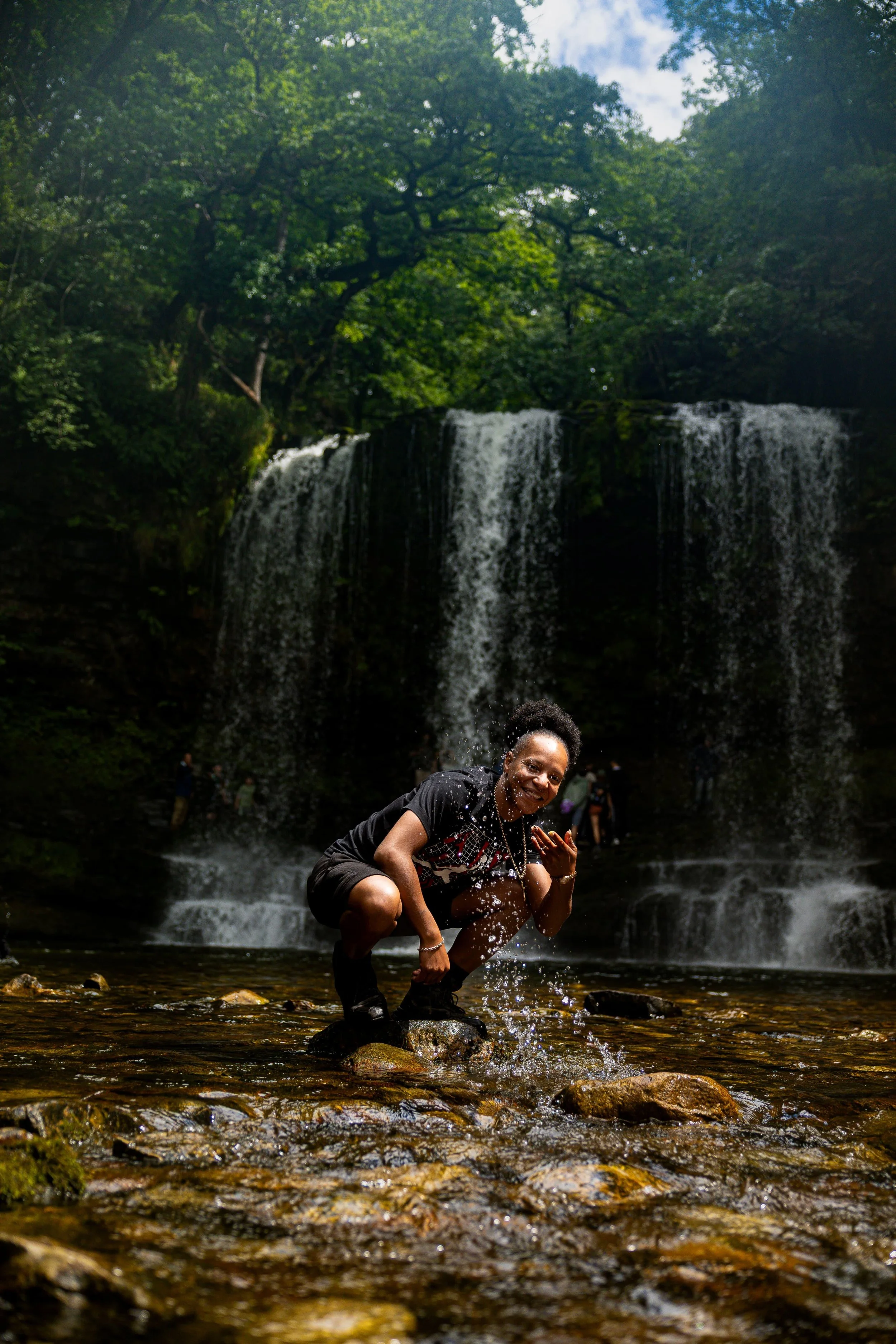 A woman crouching on rocks in a shallow stream, smiling and splashing water, with a multi-tiered waterfall and lush green trees in the background.