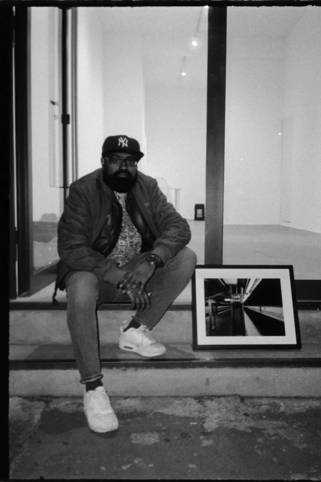 Un homme avec une casquette New York, lunettes, costume décontracté, assis devant une vitrine avec un tableau encadré affichant une photographie en noir et blanc d'un couloir ou d'une station de métro.