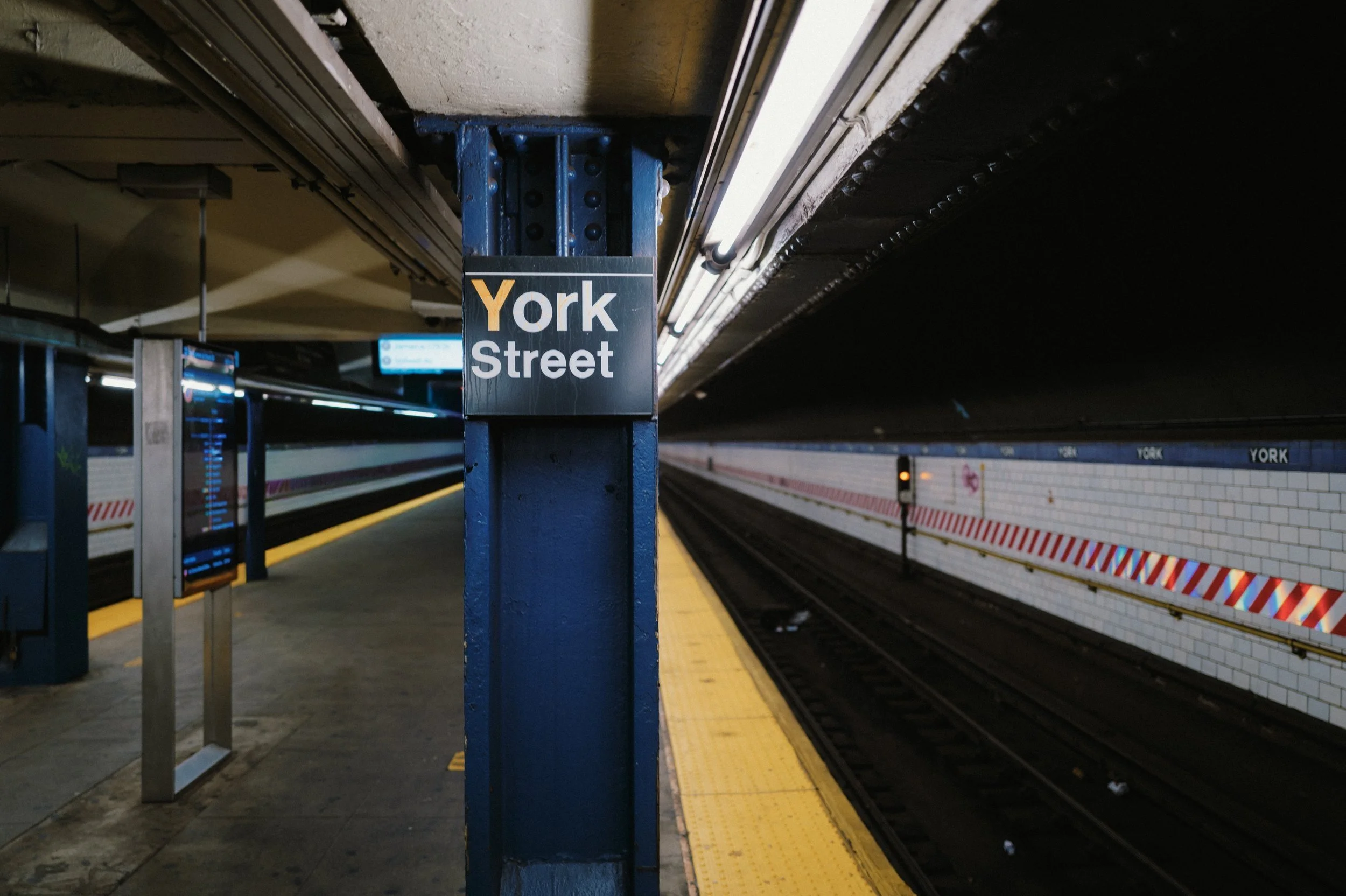 Sous-sol de métro avec panneau indiquant 'York Street', quai vide, murs en carrelage blanc, bandes de sécurité rouges et blanches, lumière fluorescent, train en arrière-plan paré à partir.