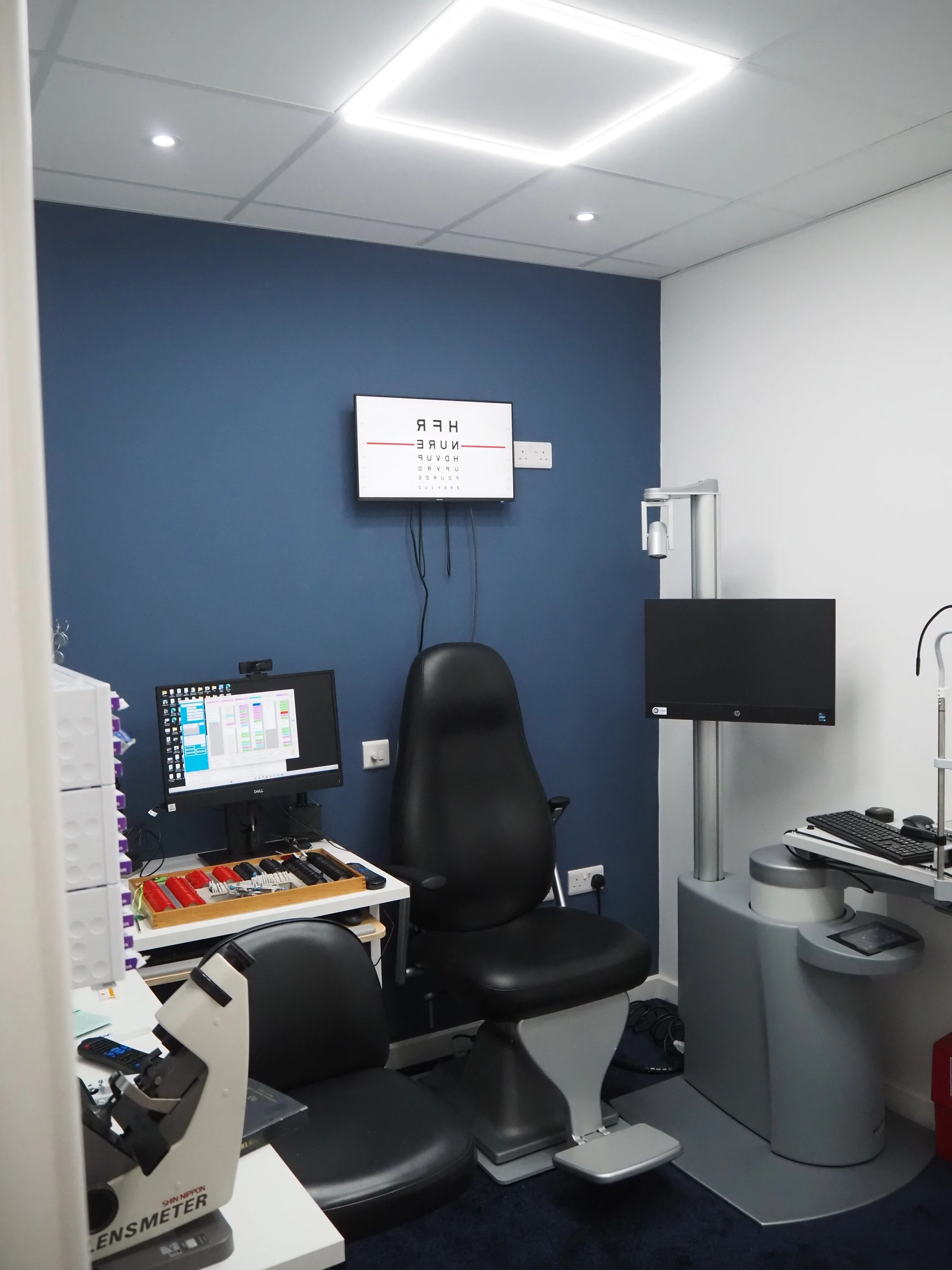 Medical examination room with equipment including a chair, monitor screens, and a vision test chart on a wall.