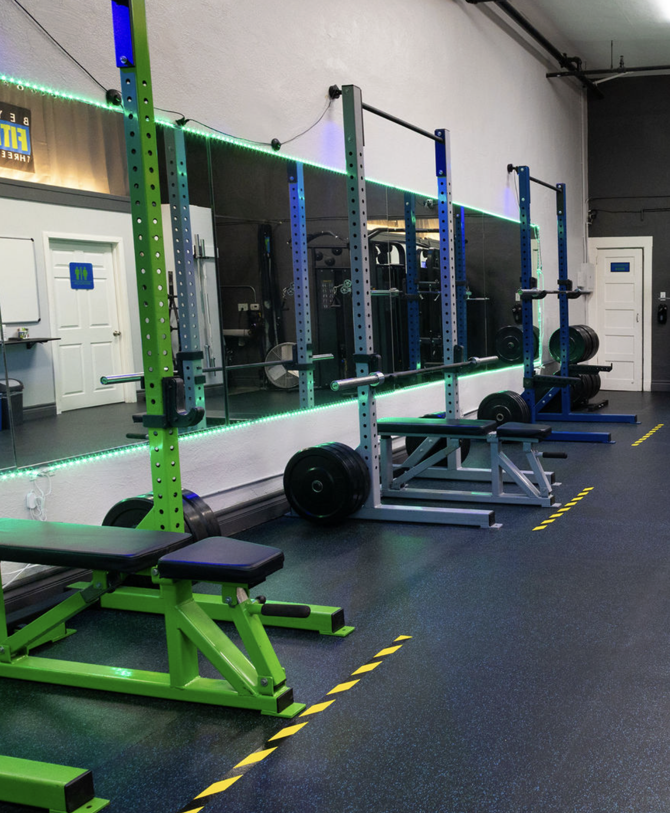An empty gym with fitness equipment including a squat rack, weight plates, benches, and a green workout machine. The gym has dark flooring, a large mirror, and LED strip lighting along the walls.