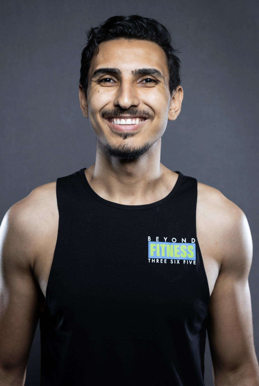 A smiling man with dark hair and a goatee, wearing a black tank top with the words 'Beyond Fitness' printed on it, standing against a gray background.