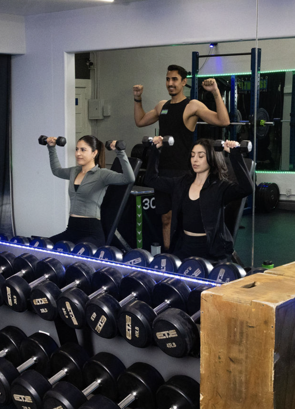 Two women sitting at a weightlifting station holding dumbbells, with a male trainer standing behind them flexing his arms, in a gym.