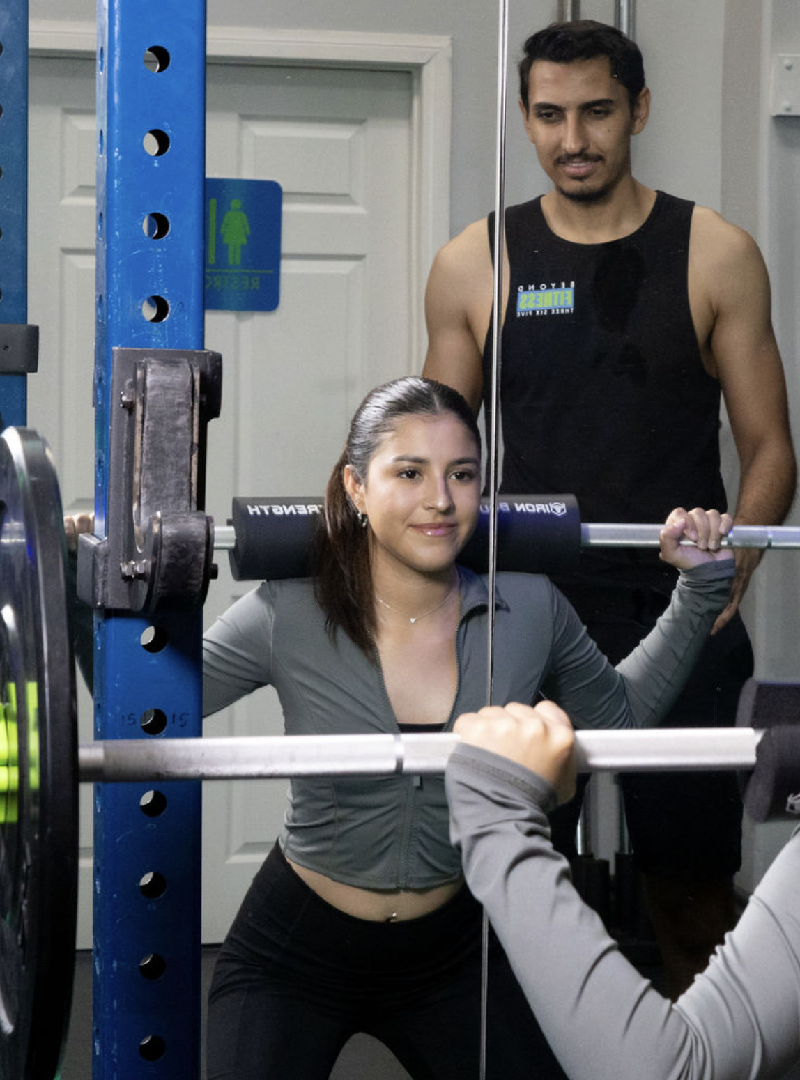 A woman in workout clothes doing squats with a barbell while a man in a black tank top watches her during exercise in a gym or fitness studio.