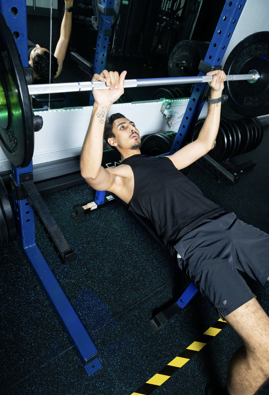 A man lying on a workout bench performing a barbell bench press at the gym, with weights on the barbell and a mirror behind him.